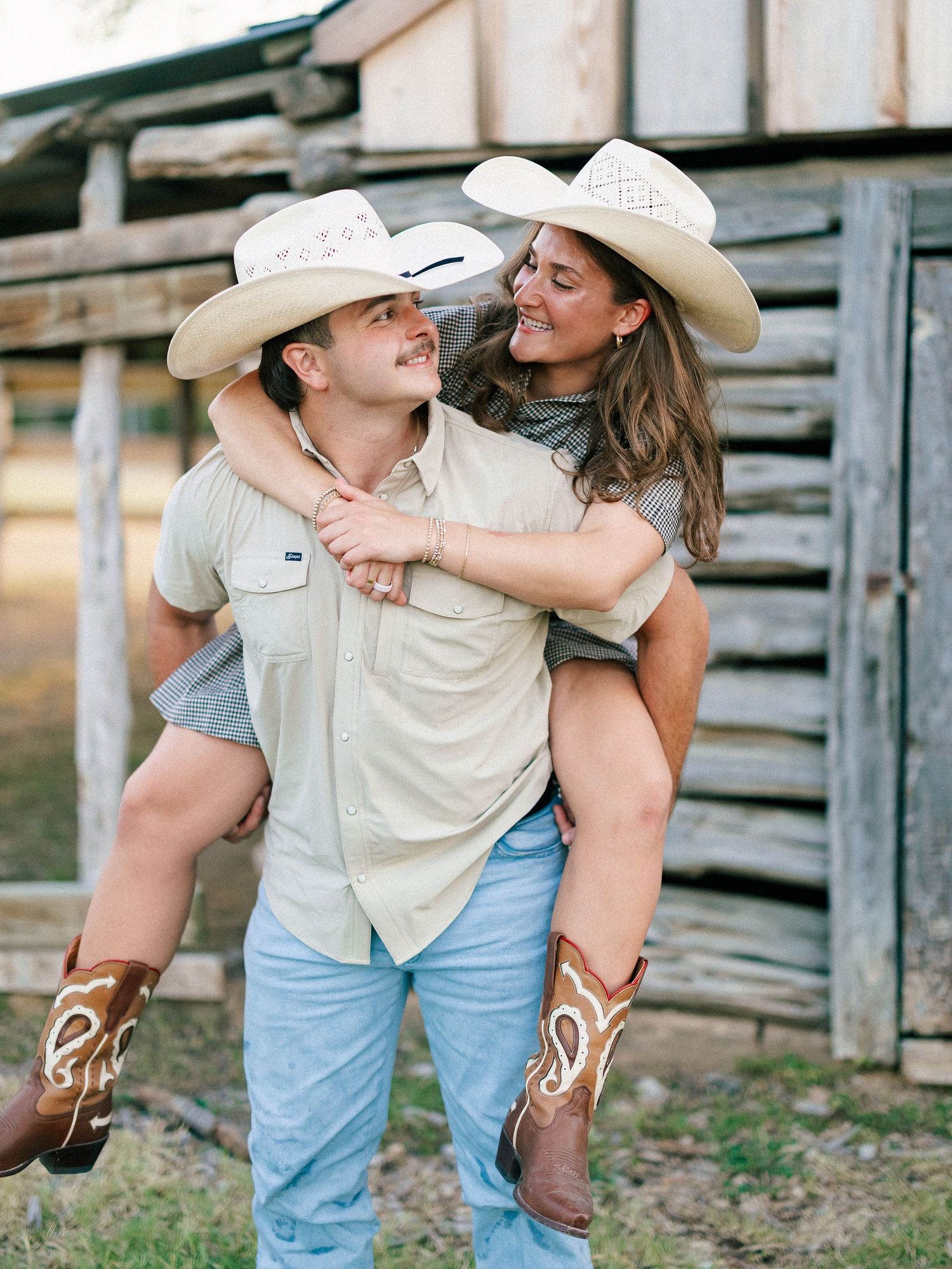 A smiling couple in matching cowboy hats and boots, with the man giving the woman a piggyback ride outdoors.
