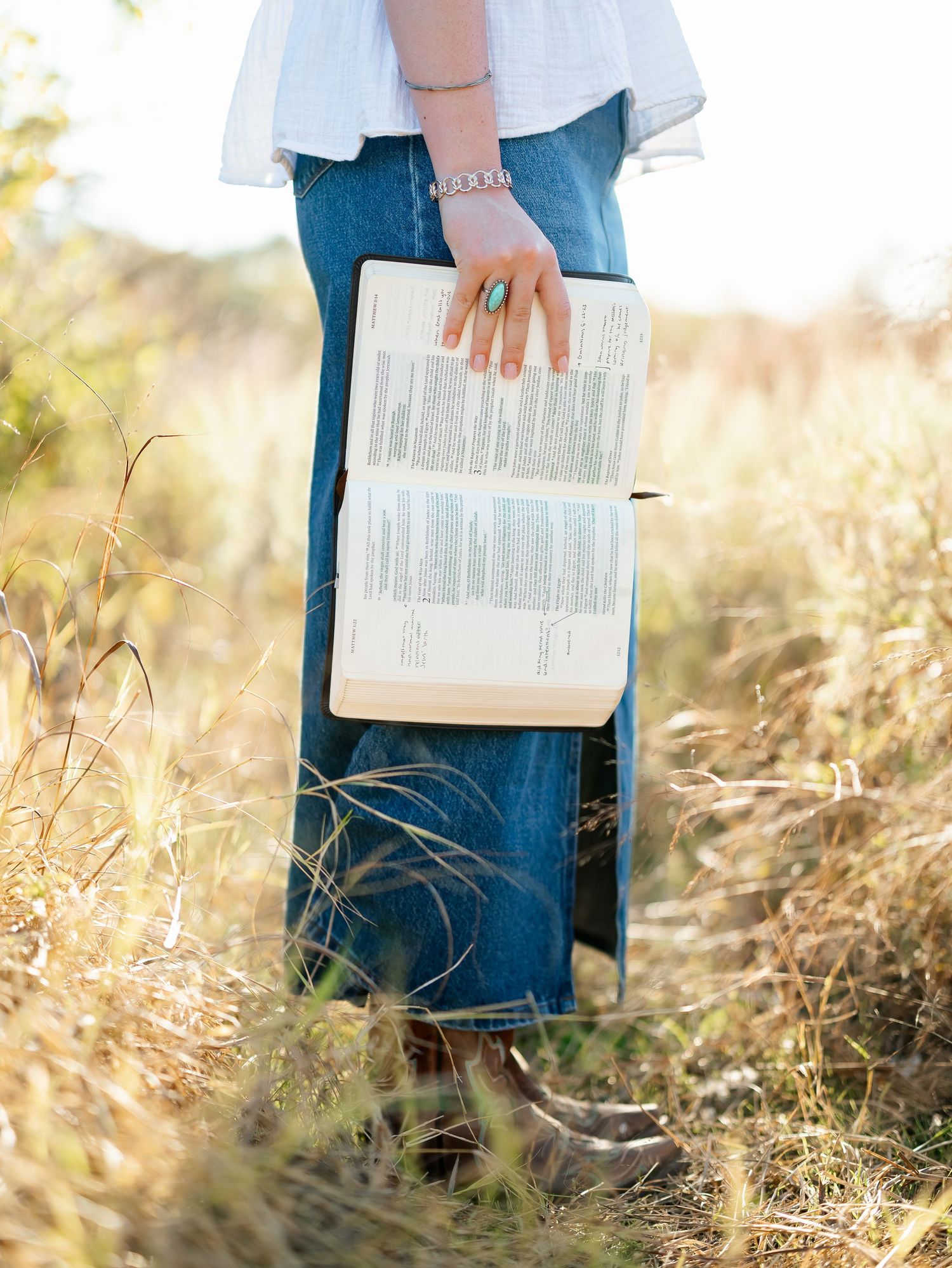 Woman in jeans holding open Bible in golden field, wearing turquoise ring and bracelet, white top, brown boots.