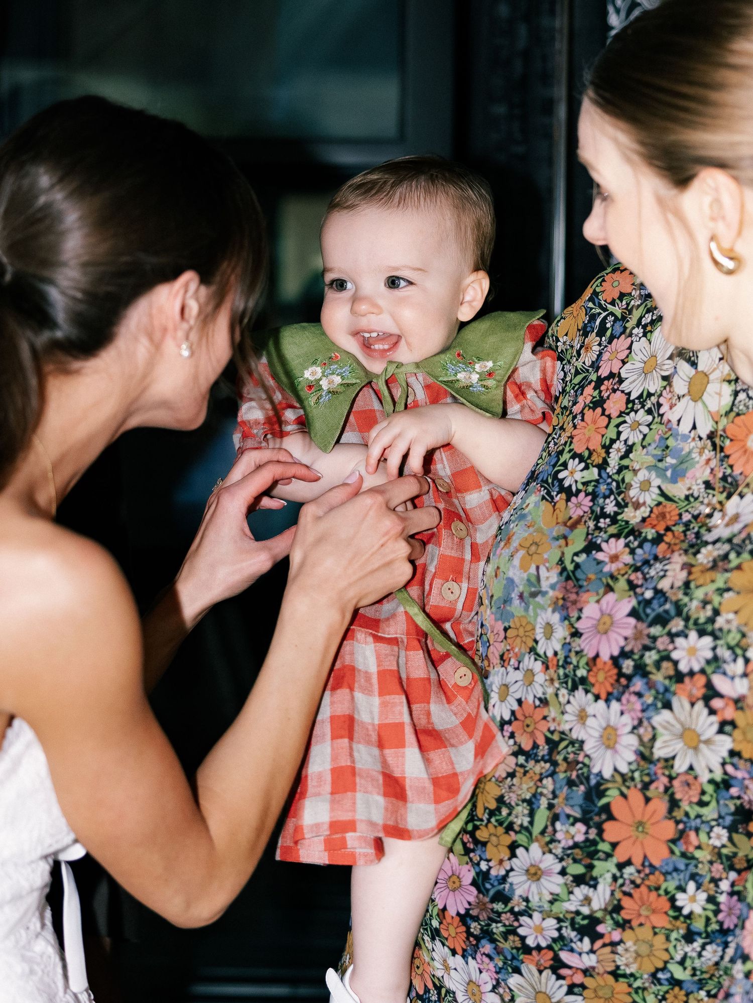 A smiling baby in red gingham dress with green collar held by woman in floral dress, greeted by another woman.