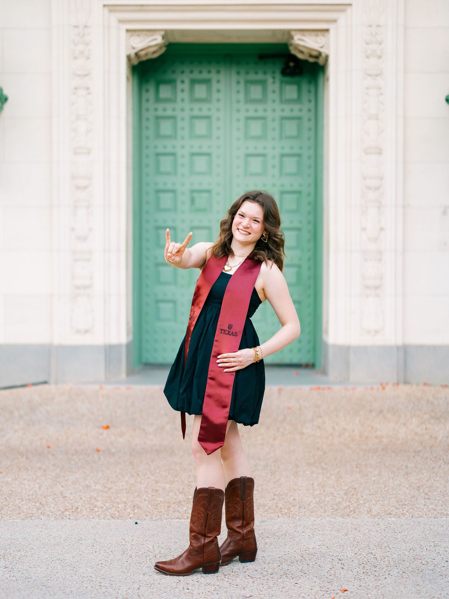 Smiling graduate in black dress with maroon stole and cowboy boots poses in front of green ornate doors.