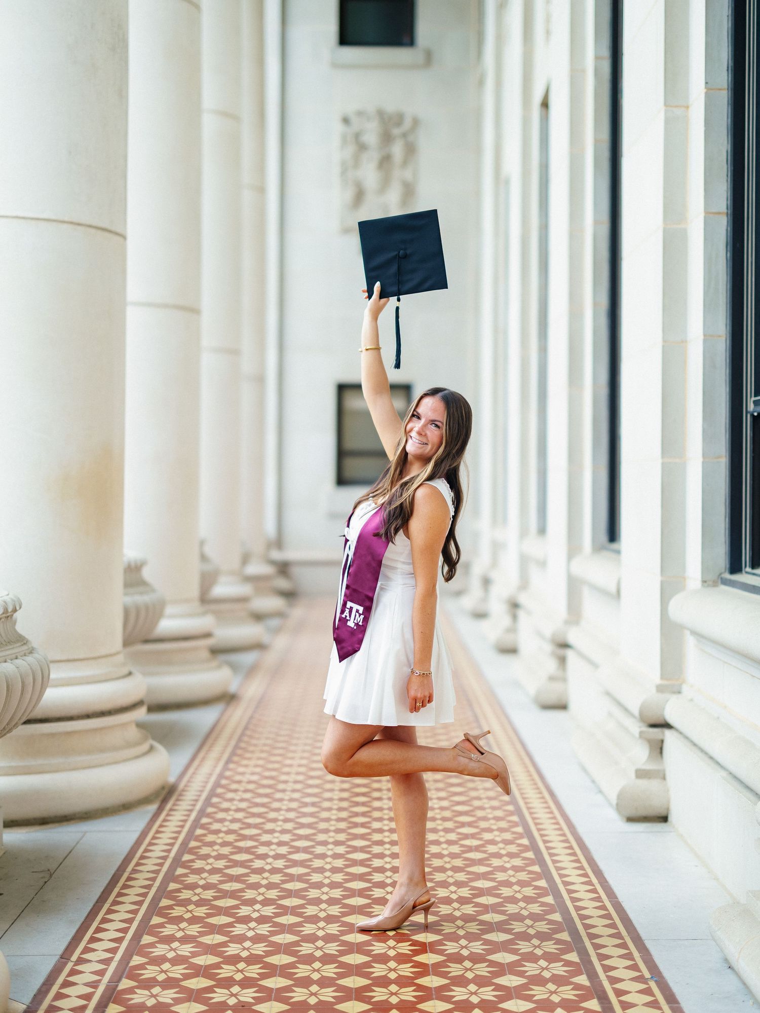 A smiling woman in a white dress holds her graduation cap aloft, wearing a maroon Texas A&M sash in a columned hall.