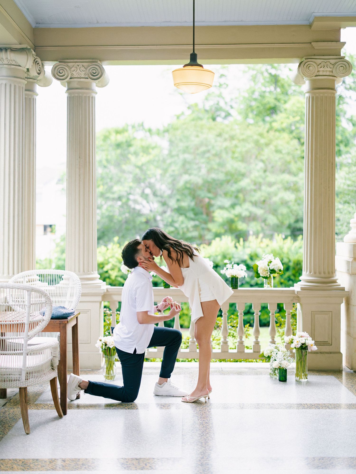 A man proposes on one knee to a woman on an elegant columned porch surrounded by floral arrangements.