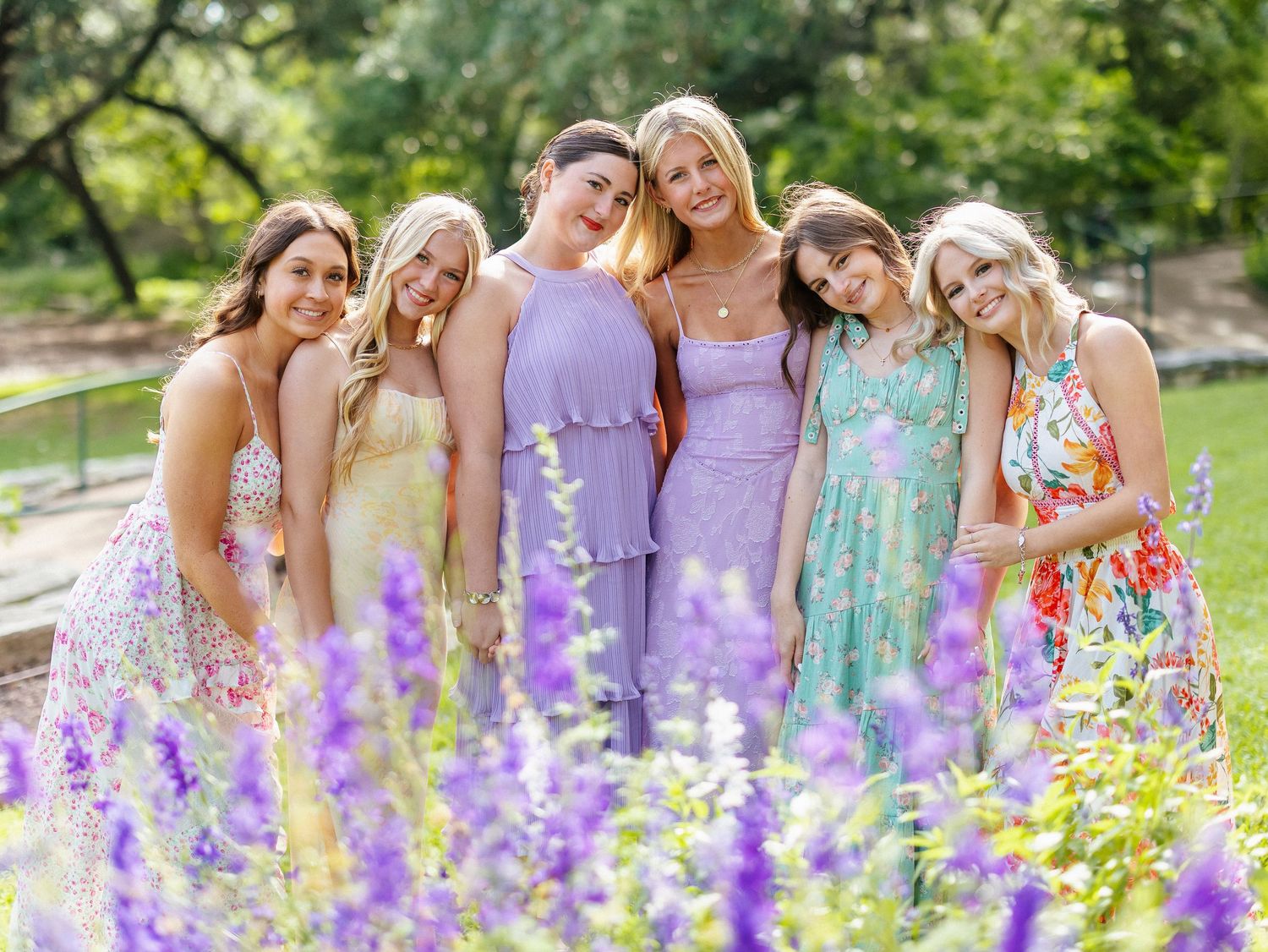 Six young women in colorful floral and pastel dresses posing together outdoors among purple wildflowers in a garden.