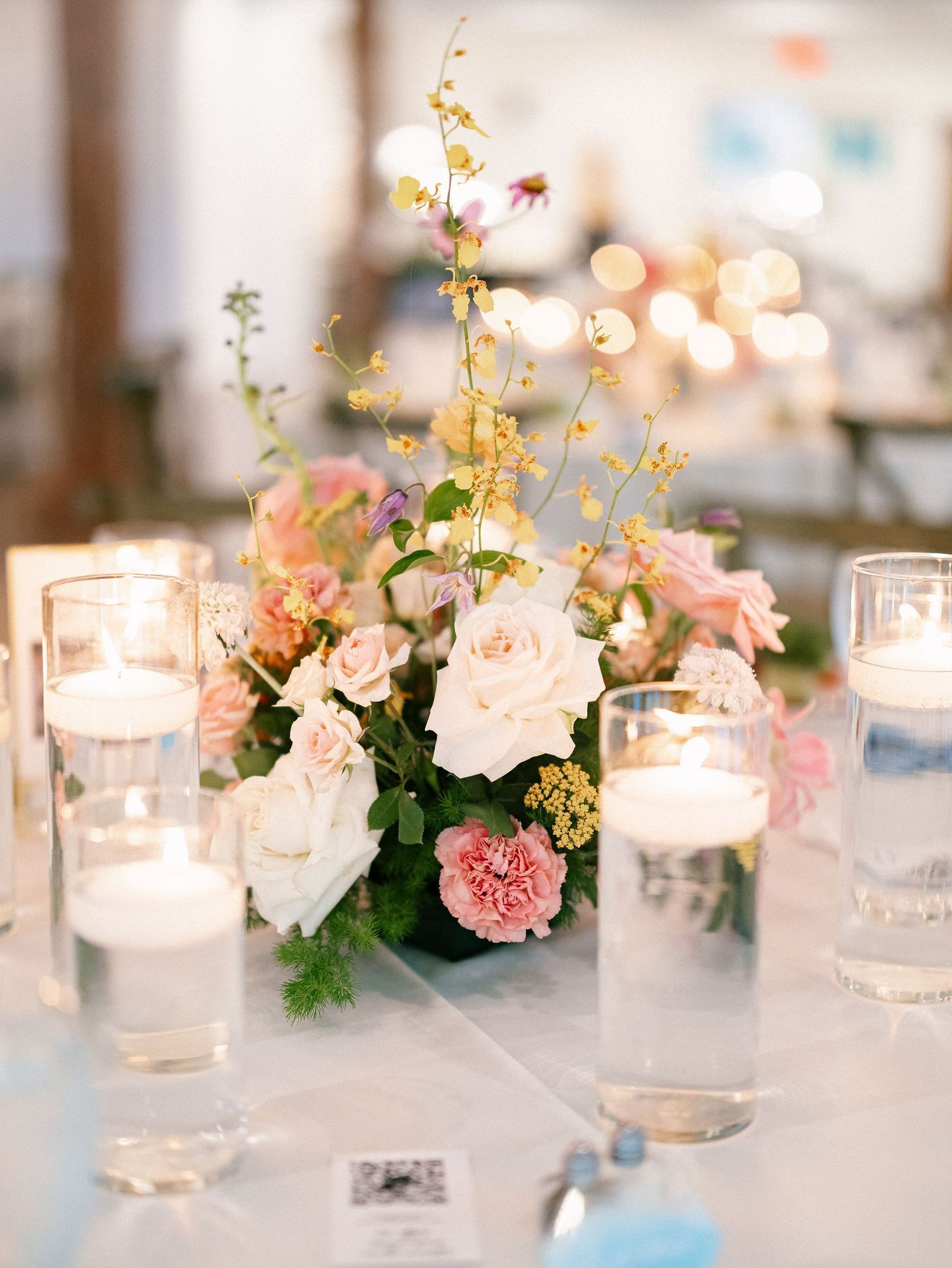 Elegant wedding centerpiece with pink and white roses, floating candles in glass cylinders, and yellow wildflowers on white linen.
