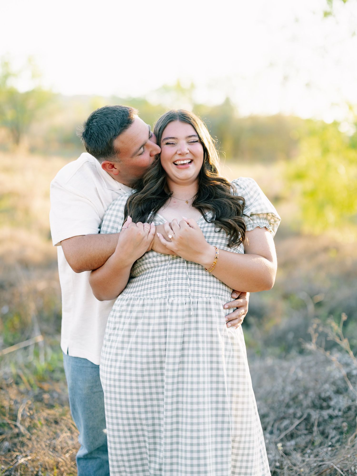 Happy couple embracing and laughing together in sun-drenched meadow during engagement shoot.