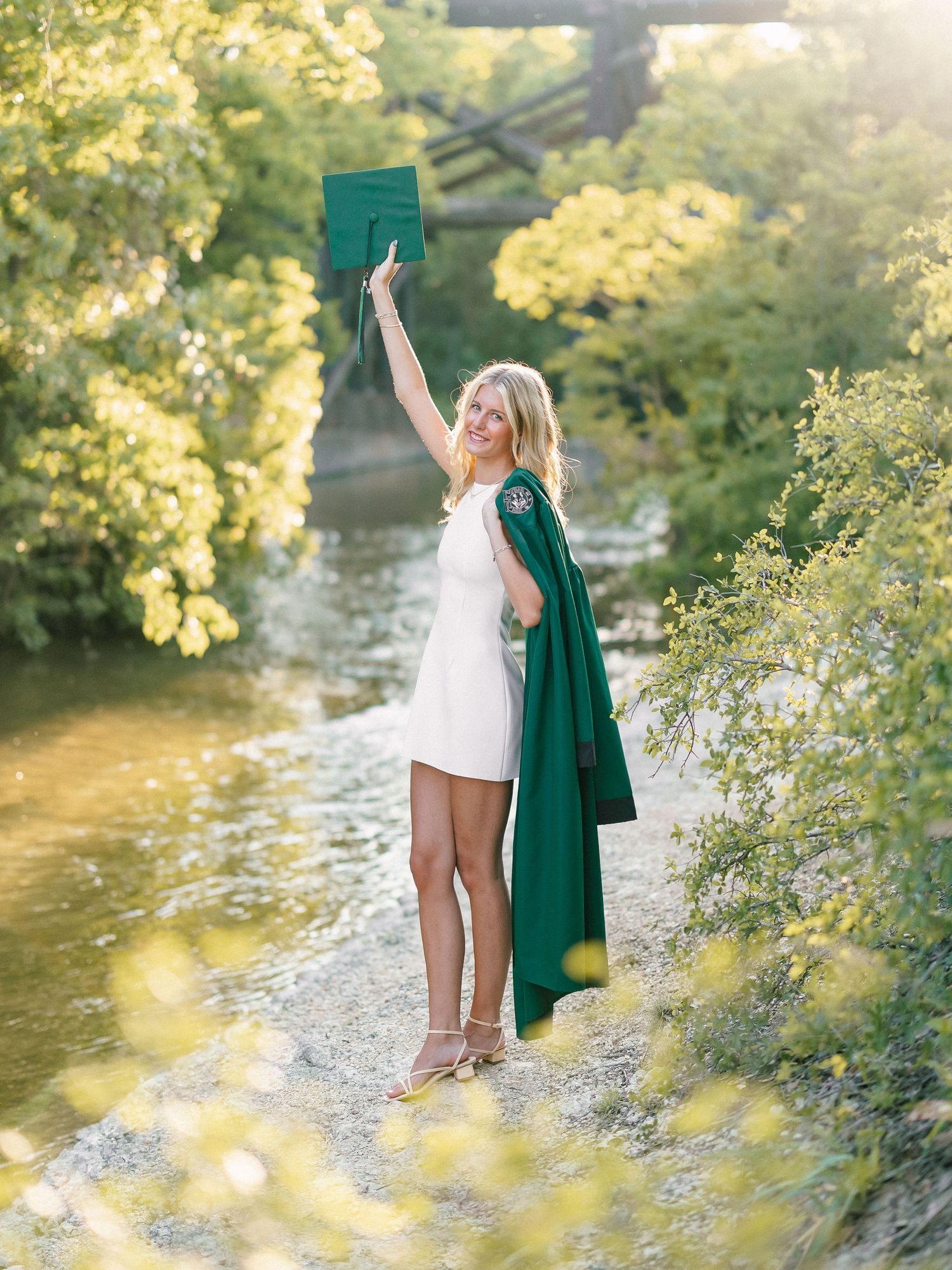 Blonde graduate celebrating in white dress and green graduation gown by a river.