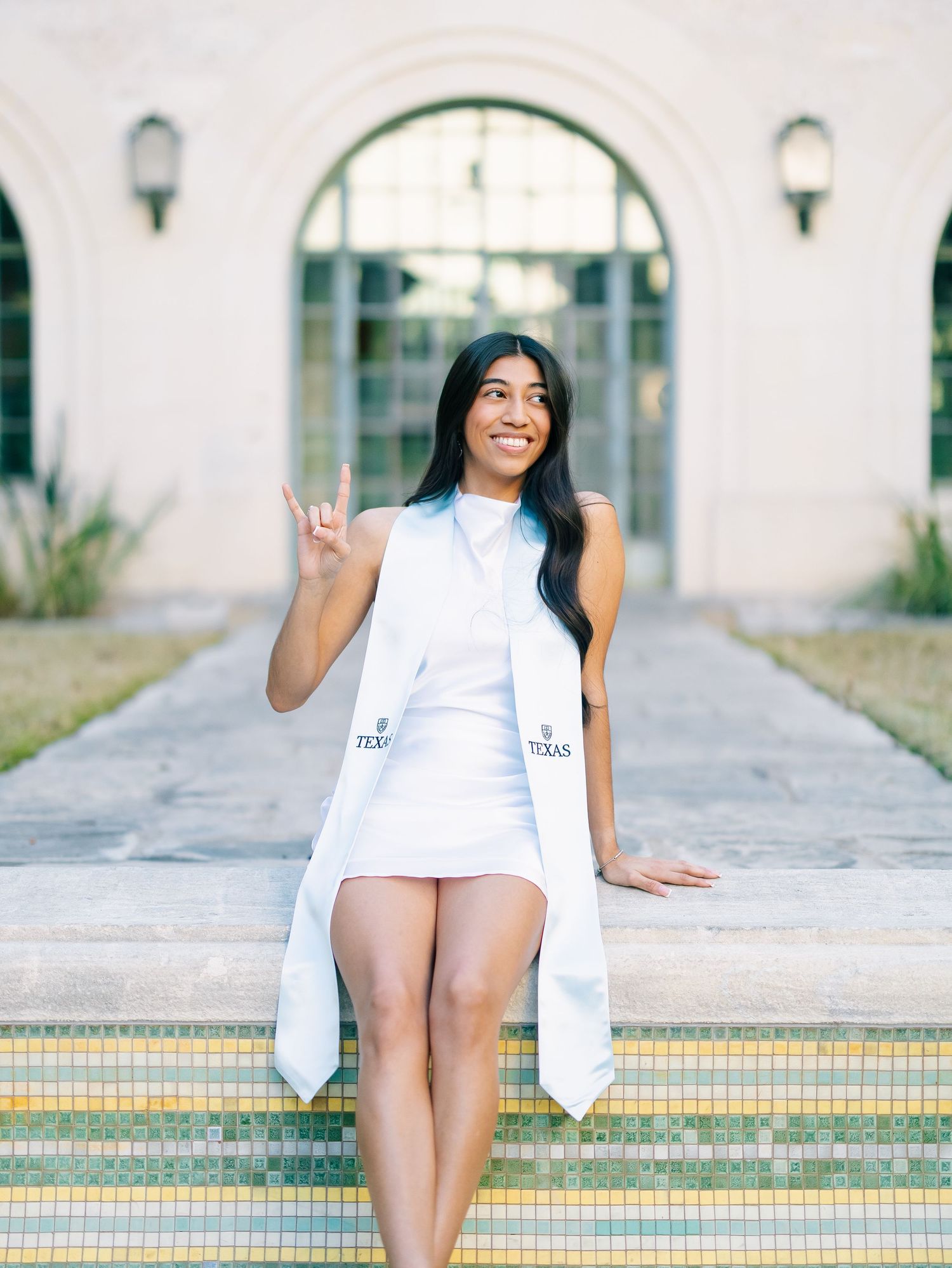 Graduate in white dress and stole makes Longhorns hand sign at University of Texas campus.