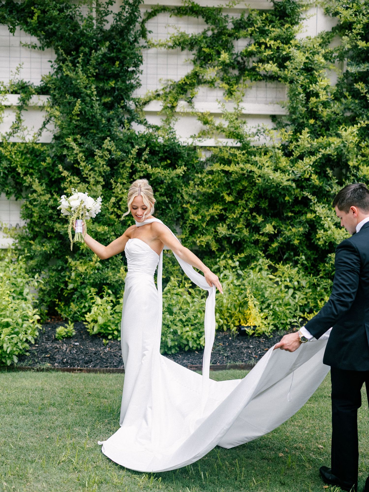 Bride in strapless white gown holds bouquet while groom spreads her train on green lawn.