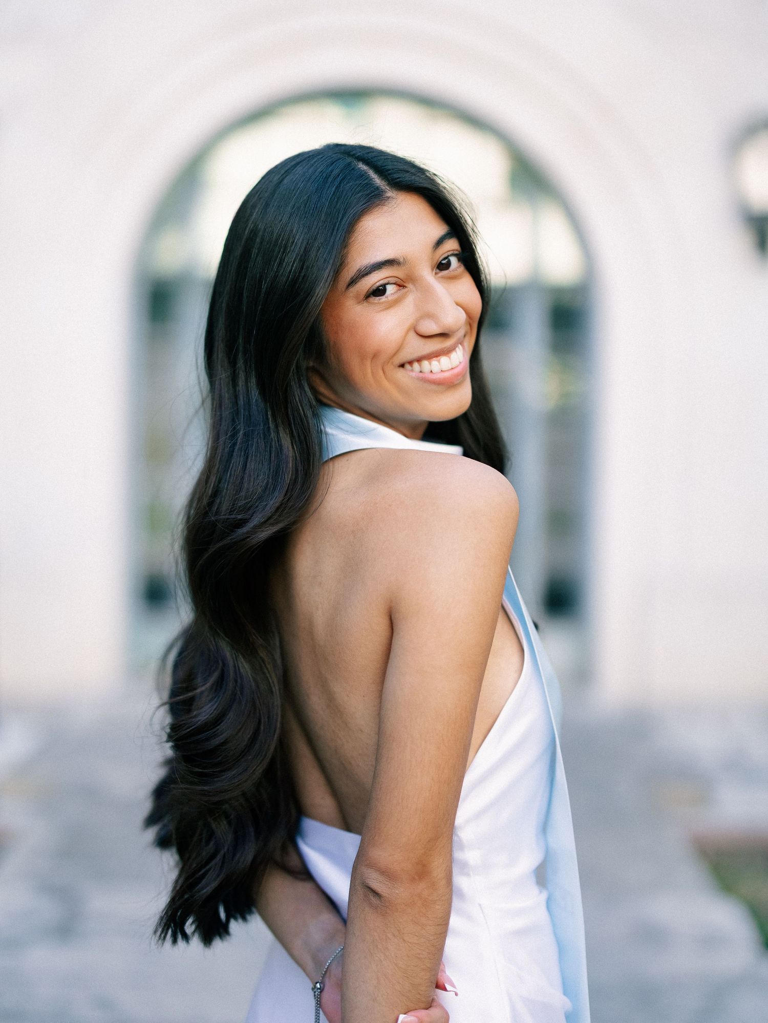 Smiling young woman with long dark hair in a white backless dress poses outdoors near an arched building.