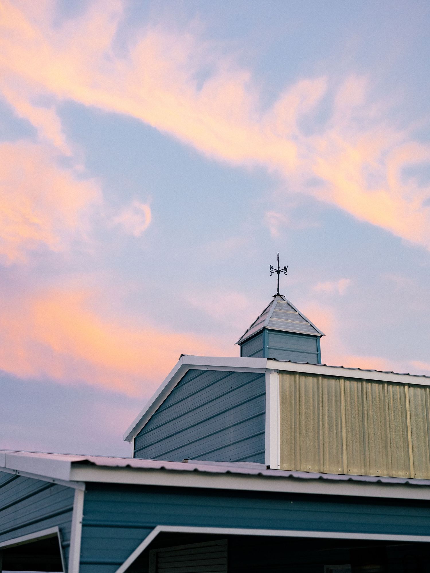 Blue barn rooftop with weather vane against a pastel pink and purple sunset sky.