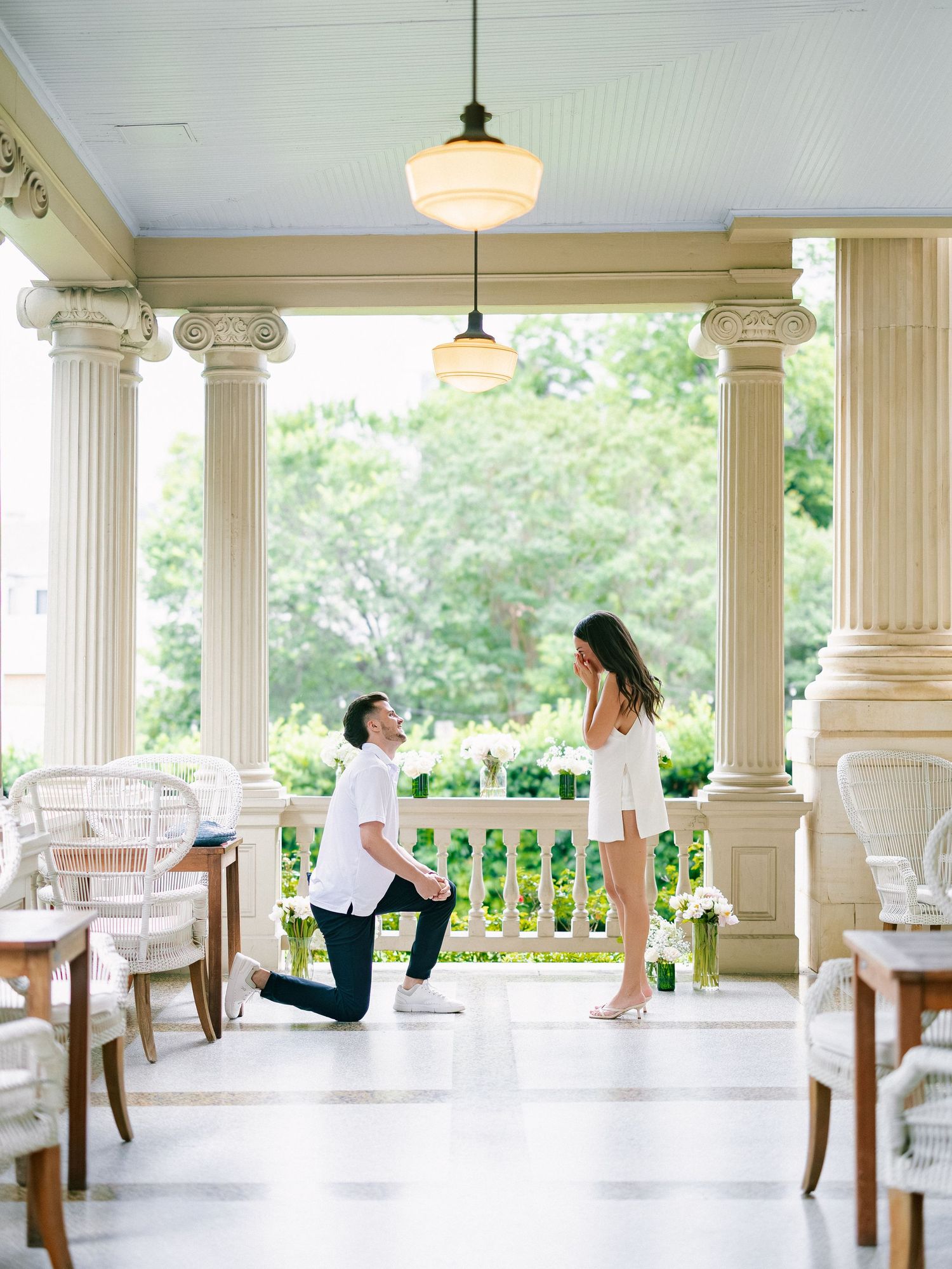 Man proposes on one knee to woman on elegant columned porch with pendant lights and white wicker furniture.