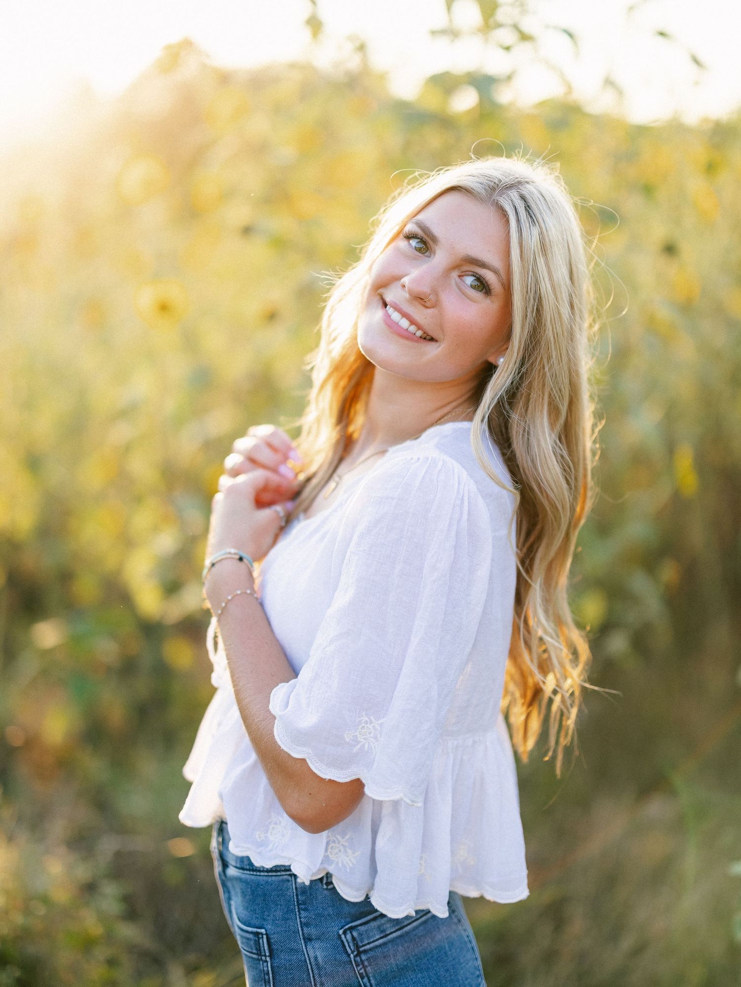 Smiling blonde woman in white eyelet top and jeans posing outdoors in a sunlit field of yellow flowers.