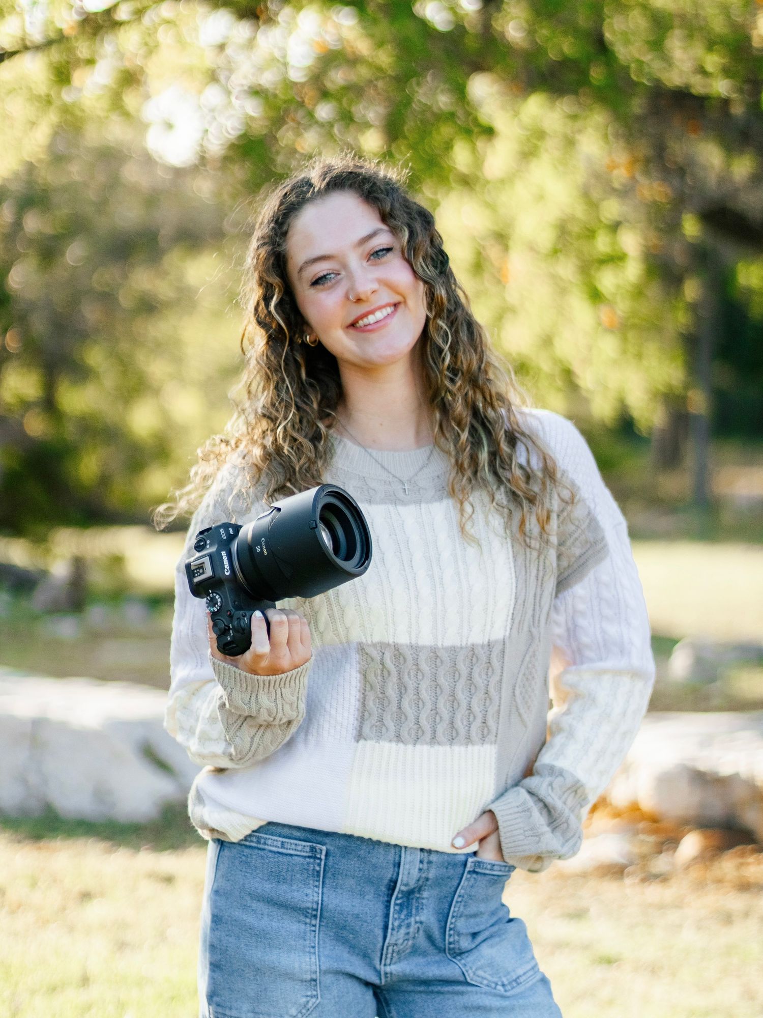 Smiling young woman with curly hair holding a DSLR camera outdoors in a park wearing a cozy sweater and jeans.