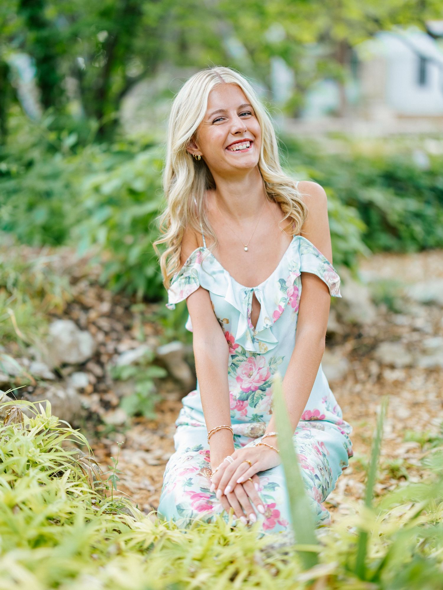 Smiling blonde woman in floral off-shoulder jumpsuit kneeling in a lush green garden setting.