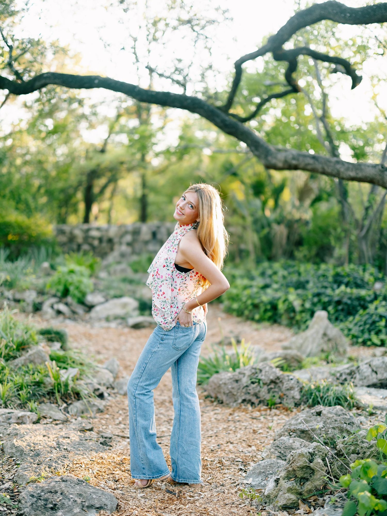 Blonde woman in floral top and flare jeans posing on rocky wooded garden path.