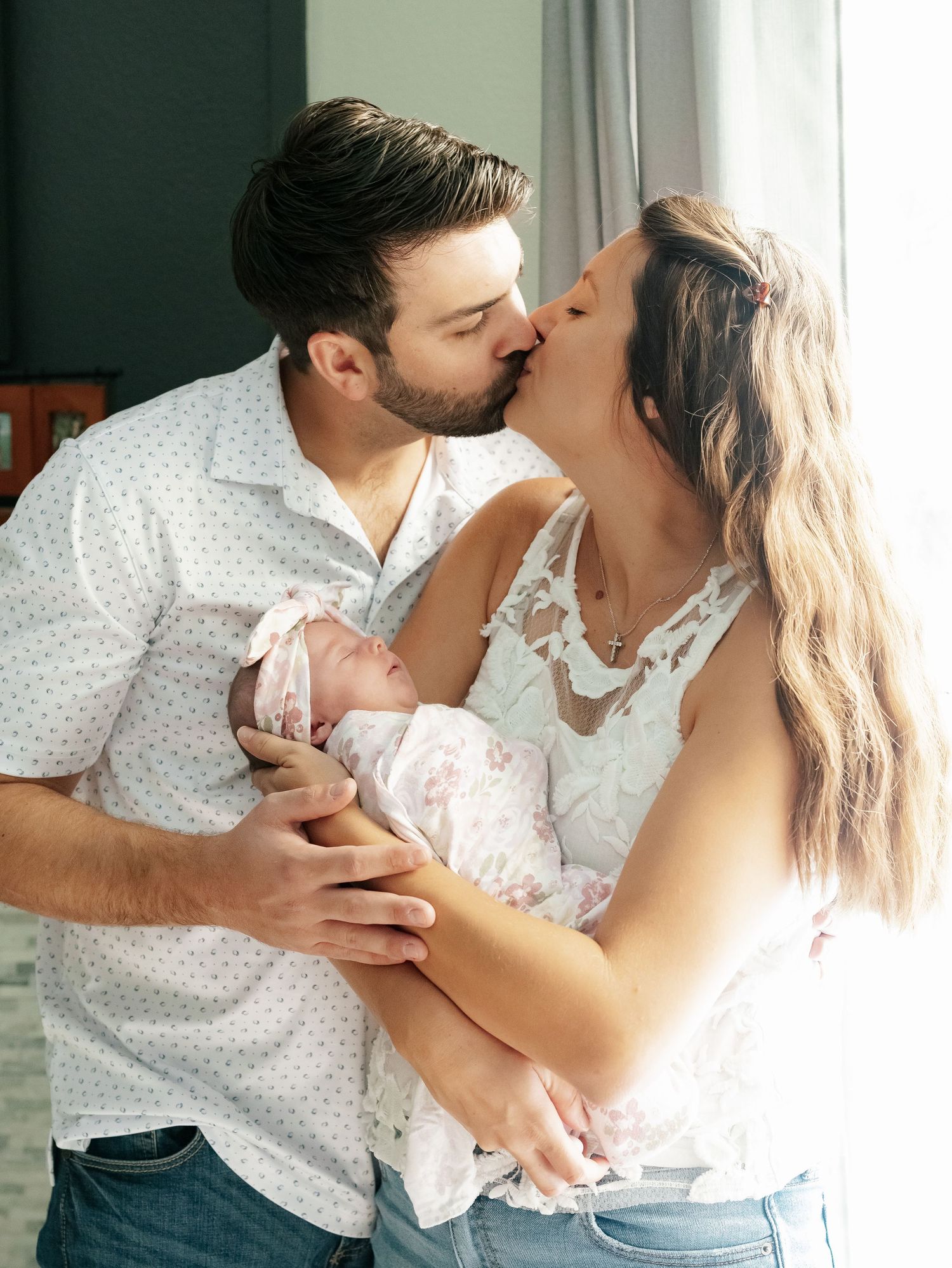 Couple kissing while holding their swaddled newborn baby together near a bright window.