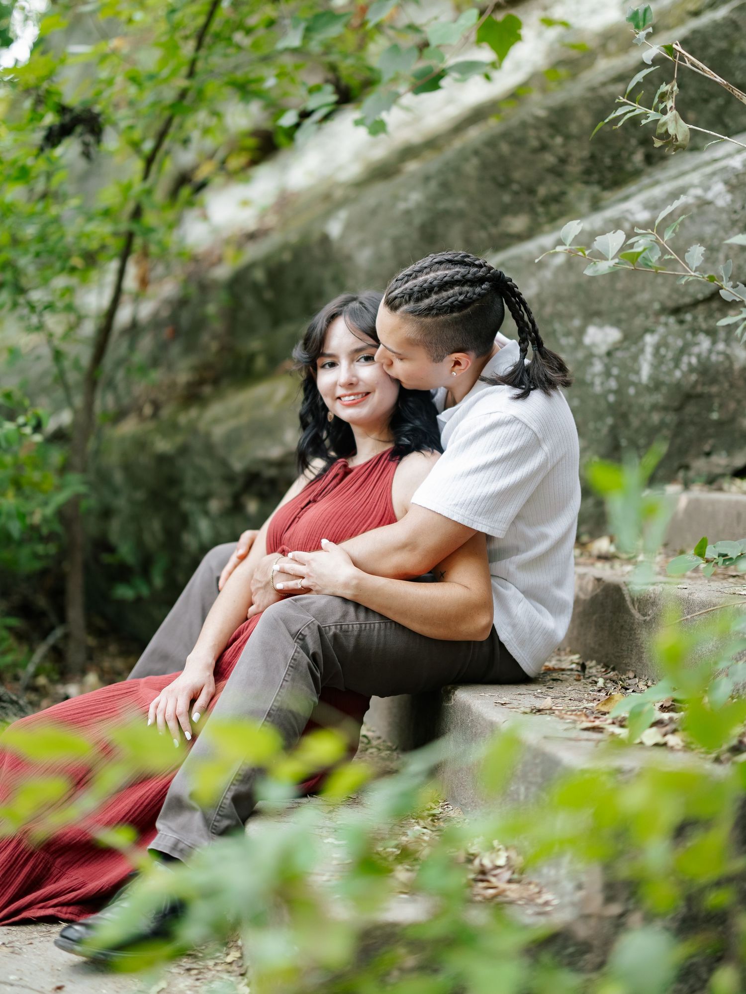 Romantic couple sitting on stone steps outdoors, man kissing woman's cheek in lush greenery.