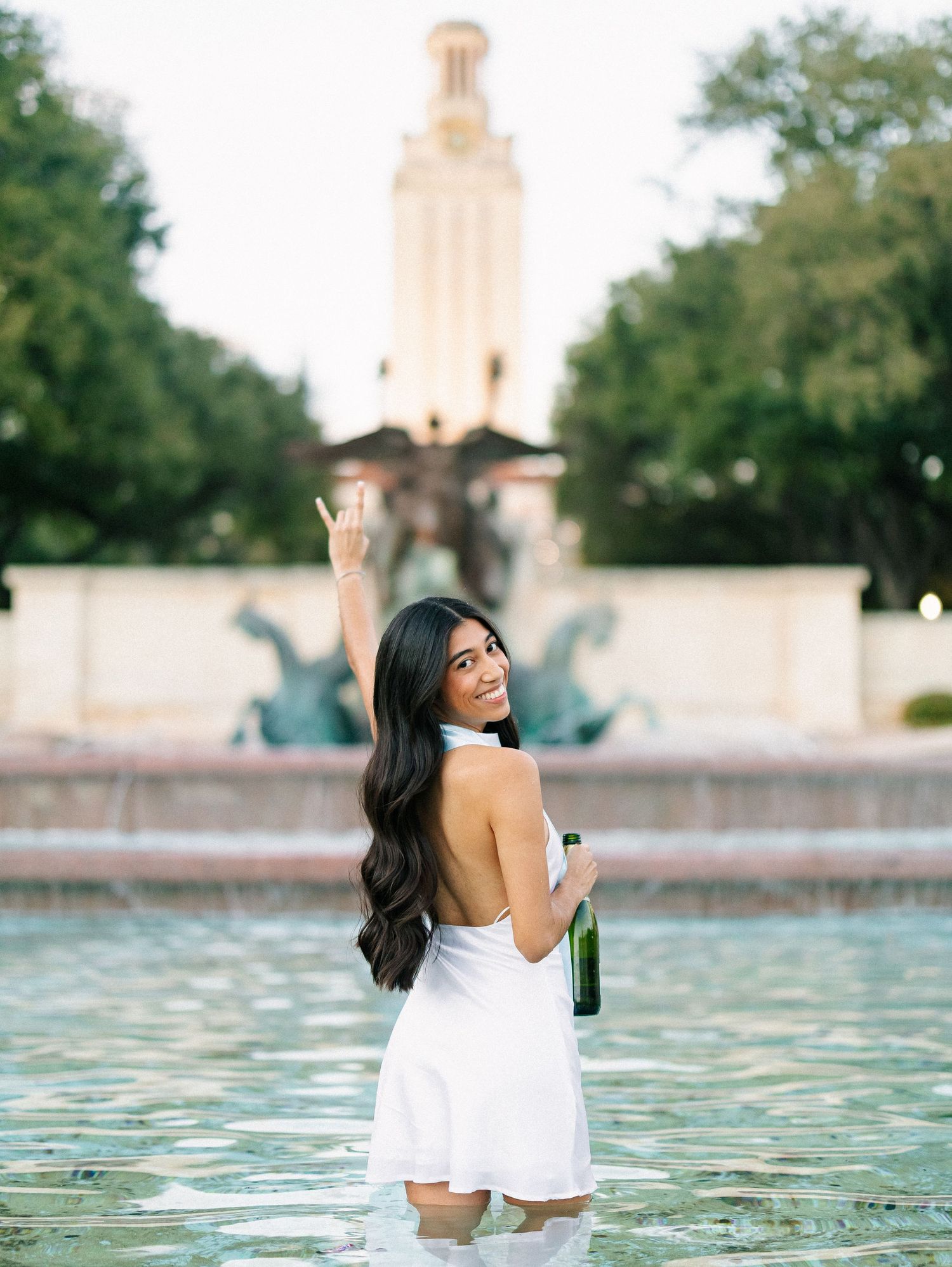 Woman in white dress celebrating graduation at UT Austin fountain, holding champagne bottle with hook em horns sign.
