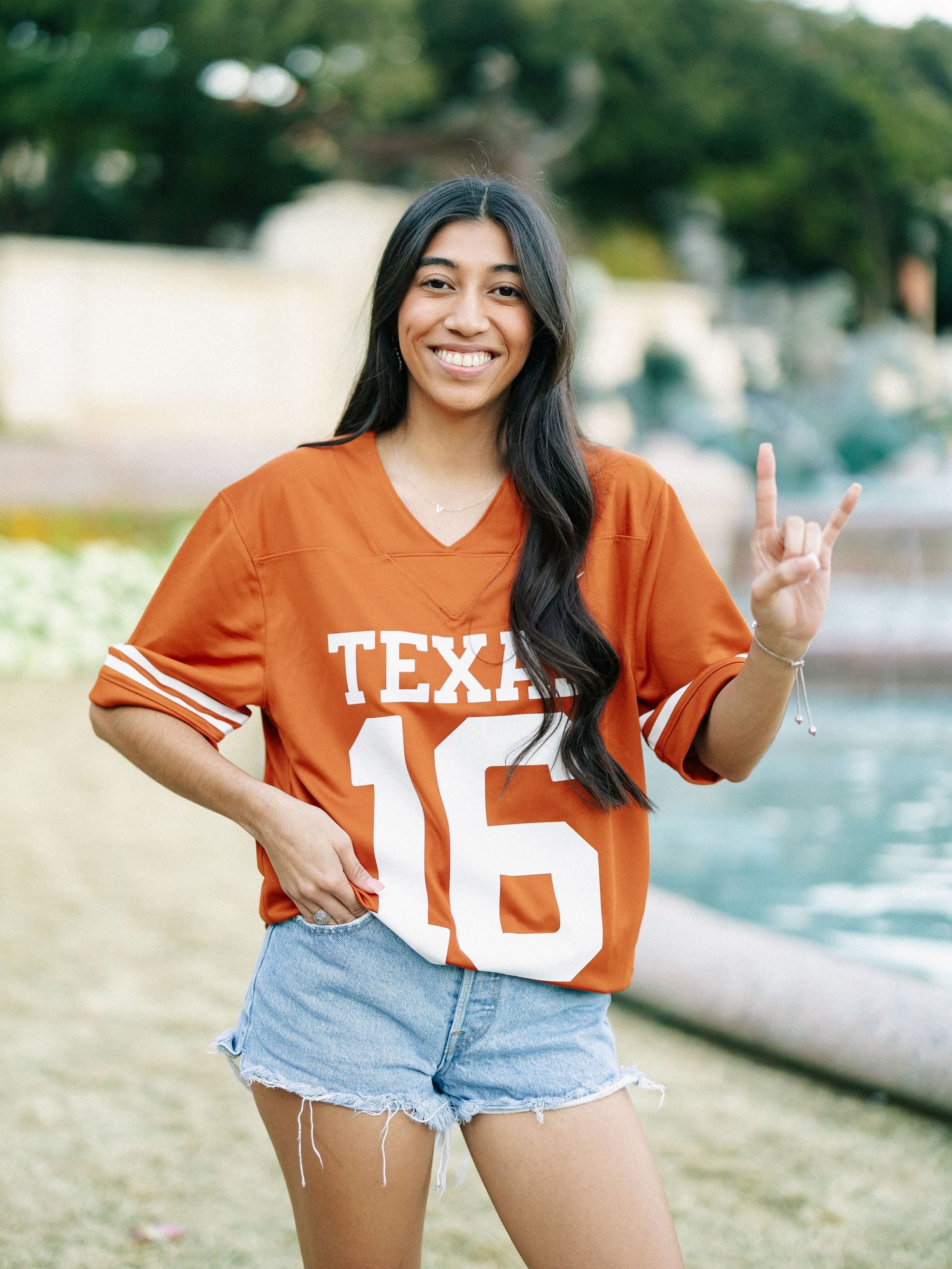 Young woman in Texas Longhorns orange jersey number 16 making hook em horns sign near fountain.