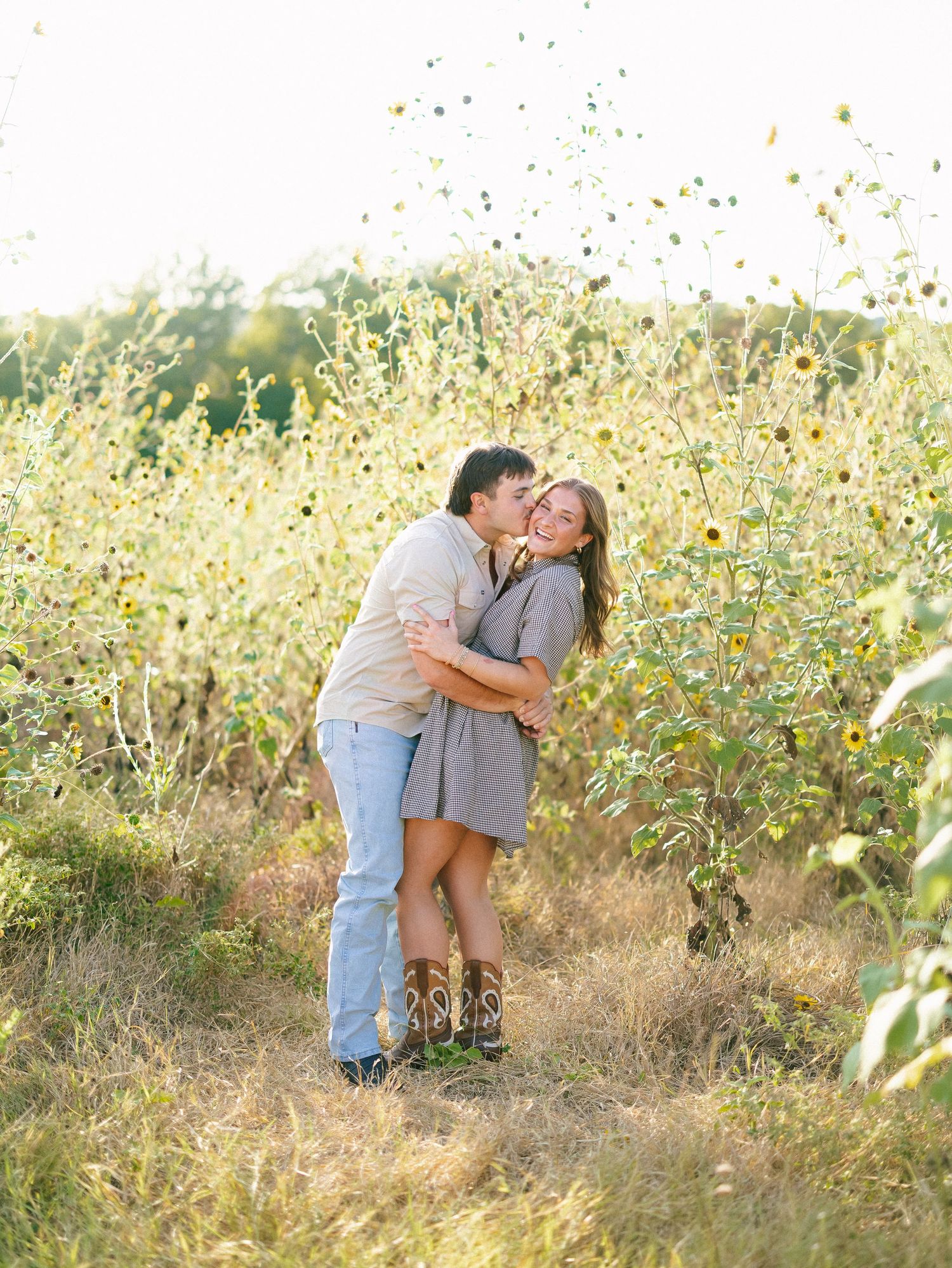 Couple embracing and kissing in a sunflower field during golden hour, woman wearing cowboy boots and striped dress.