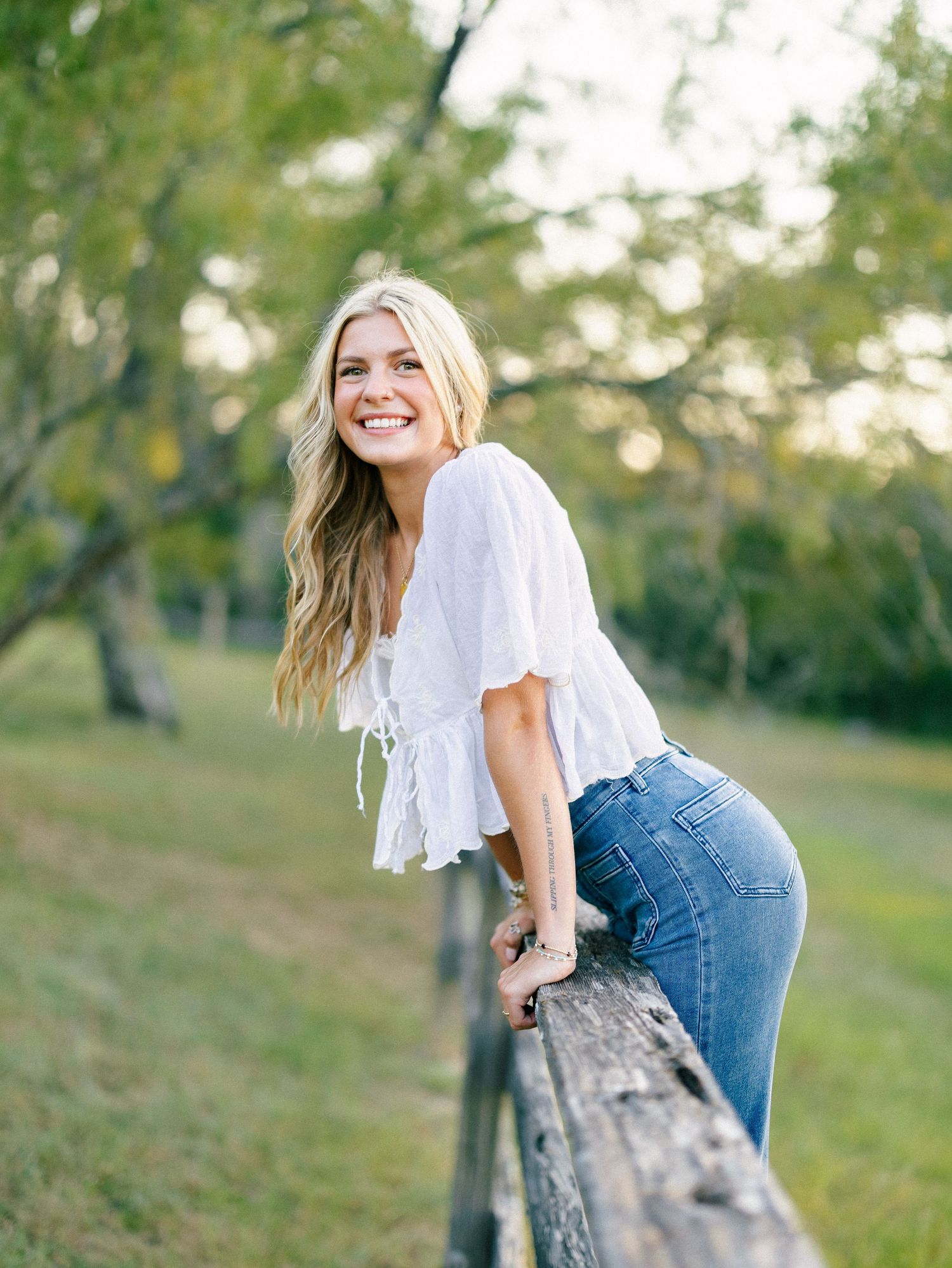Smiling young woman in white top and jeans leaning on rustic wooden fence in lush green outdoor setting.