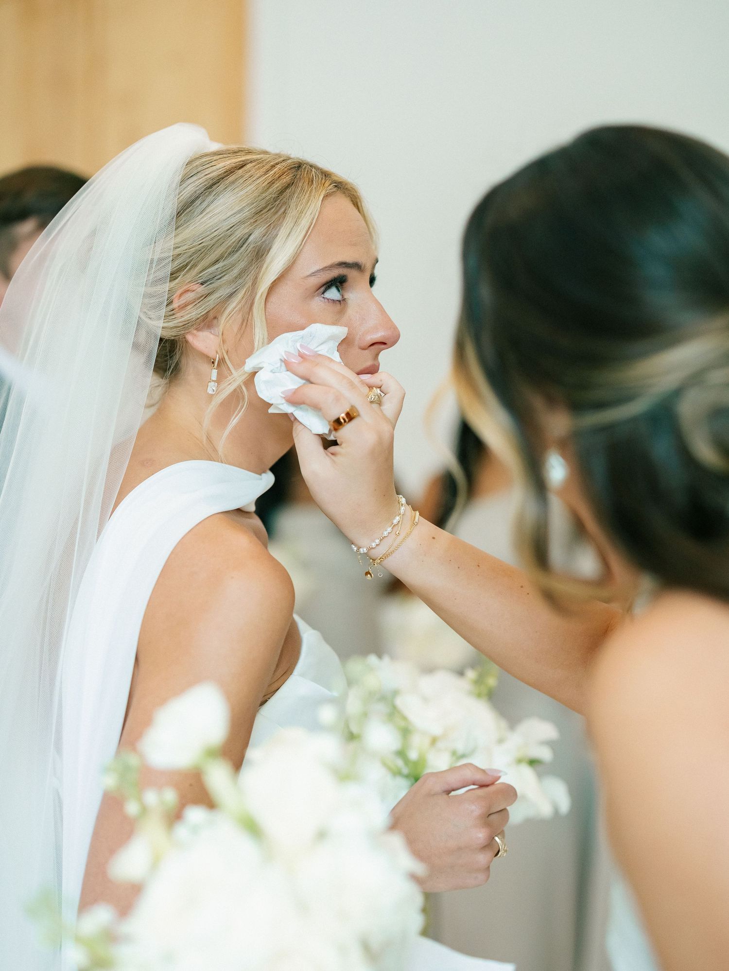 Bridesmaid gently dabbing tears from emotional bride's face before ceremony.
