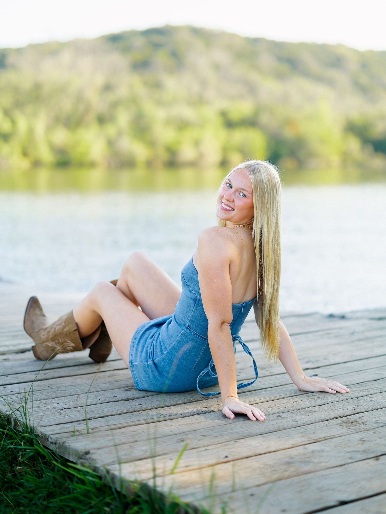 Smiling blonde woman in denim romper and cowboy boots sitting on a wooden dock by a lake.