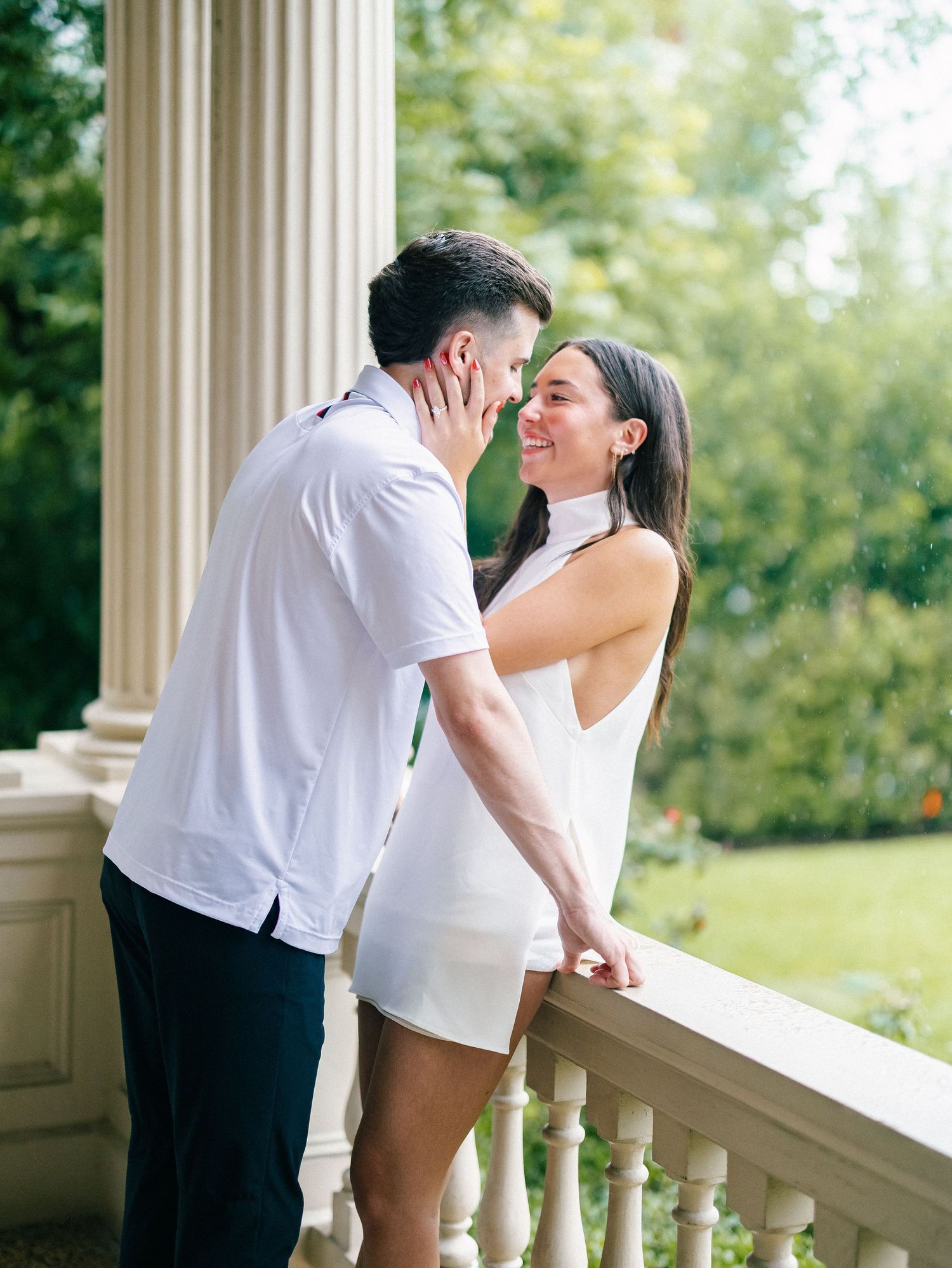 Couple in white outfits sharing a romantic moment on an elegant columned balcony surrounded by lush greenery.