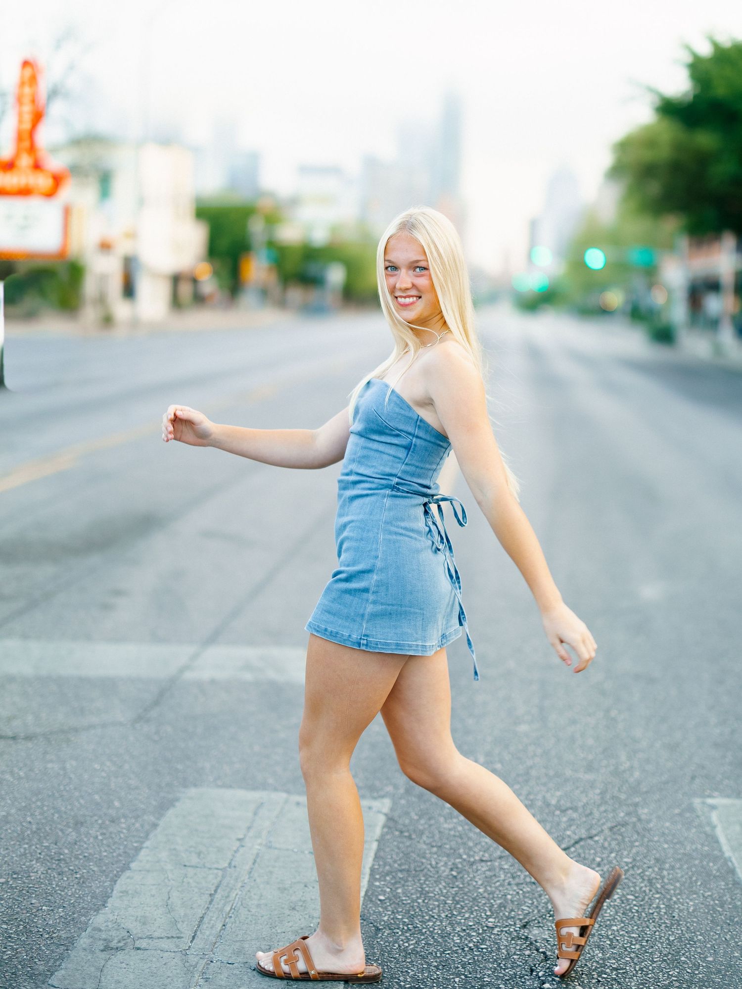 Smiling blonde woman in denim mini dress and sandals crossing a city street with urban skyline backdrop.