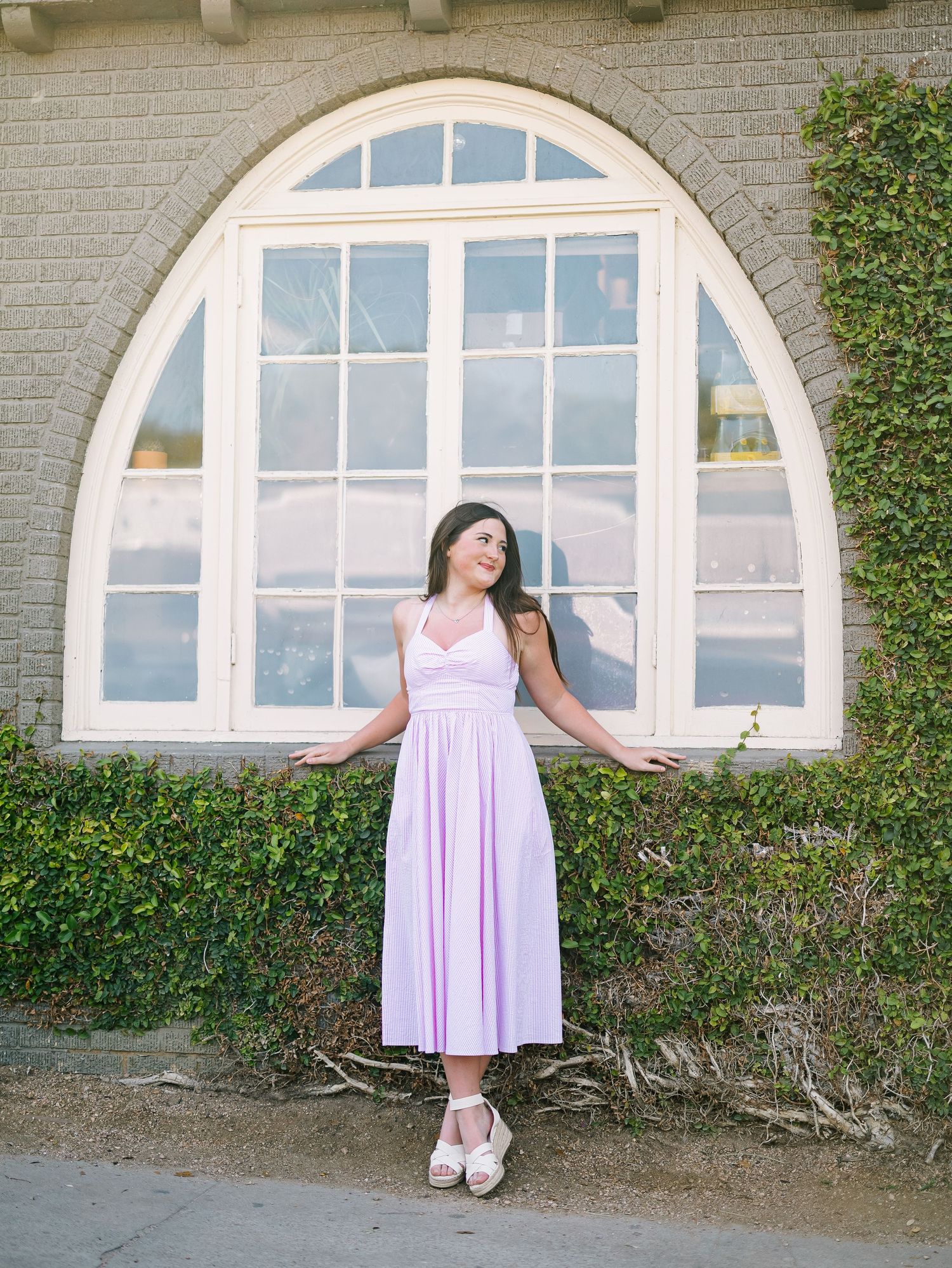 Woman in lavender midi dress posing in front of arched window with ivy-covered walls outdoors.