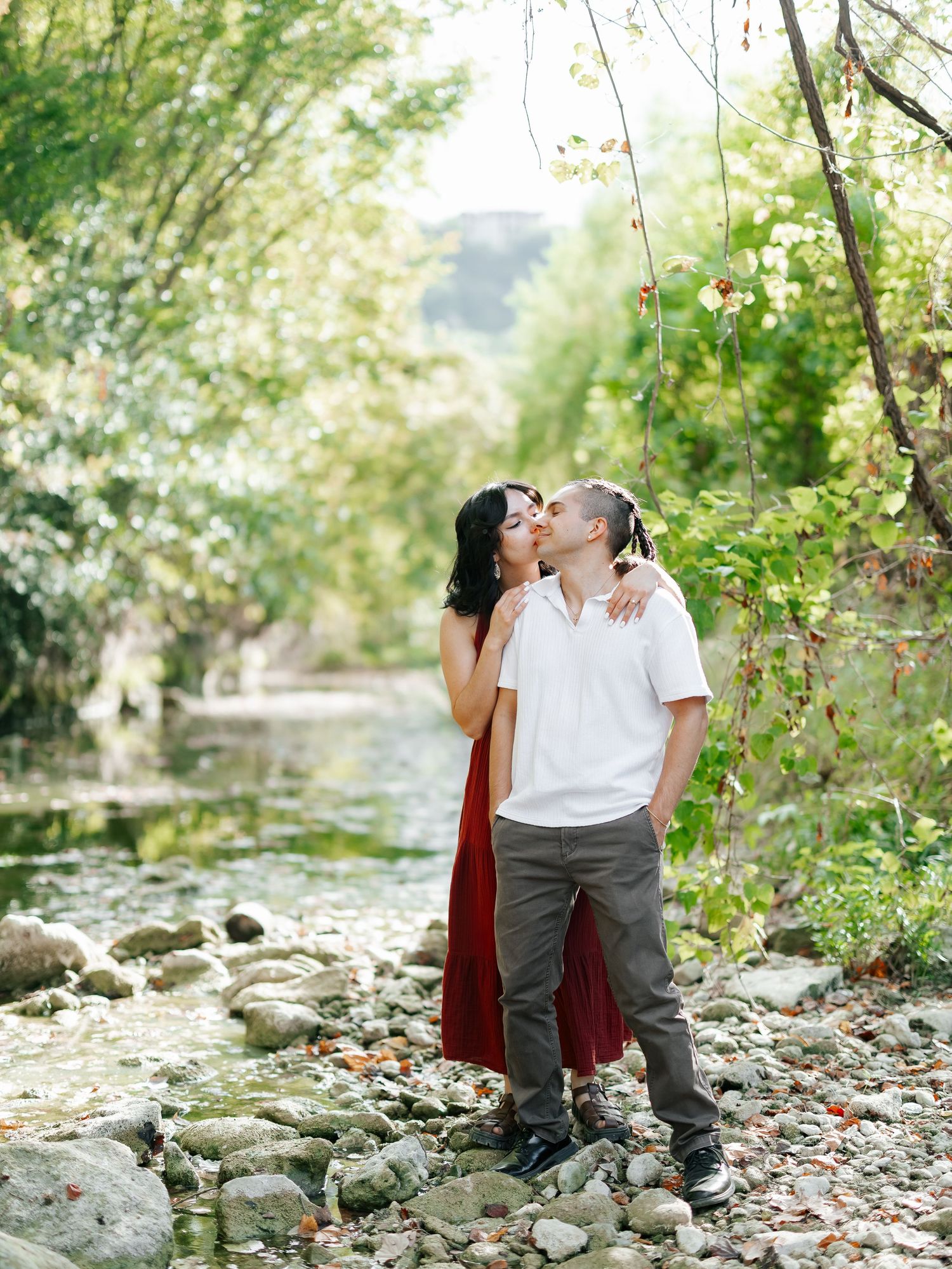 Couple sharing a kiss beside a peaceful tree-lined creek with rocky shores in natural setting.
