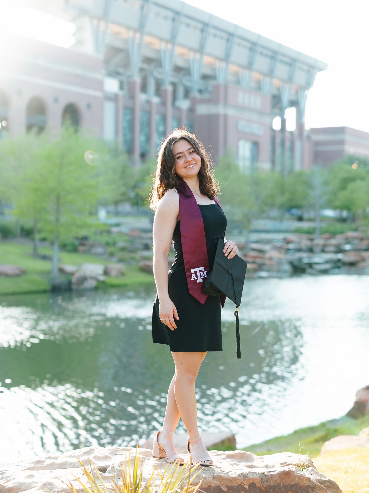 Texas A&M graduate poses in black dress with maroon sash near campus pond.