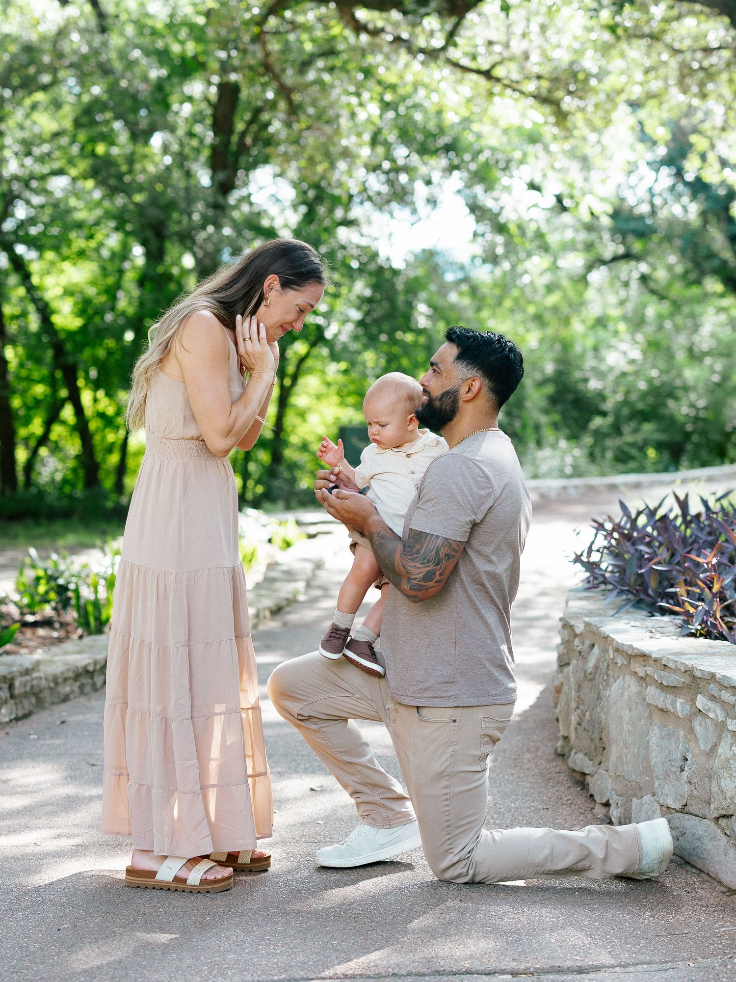 Man on one knee proposing to woman in blush dress while holding baby in lush garden setting.