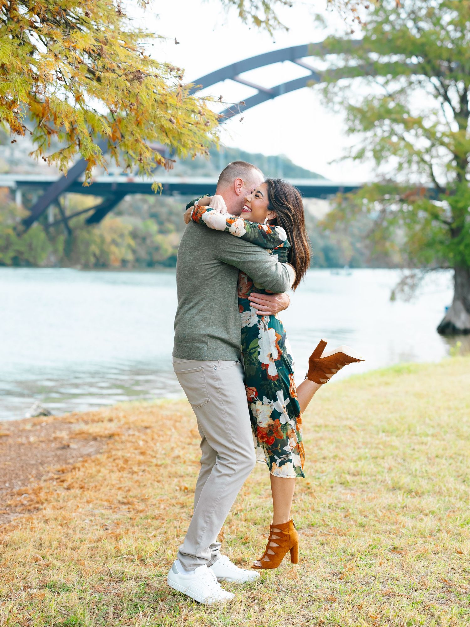 Happy couple hugging and laughing near a river with a scenic arched bridge and fall foliage.