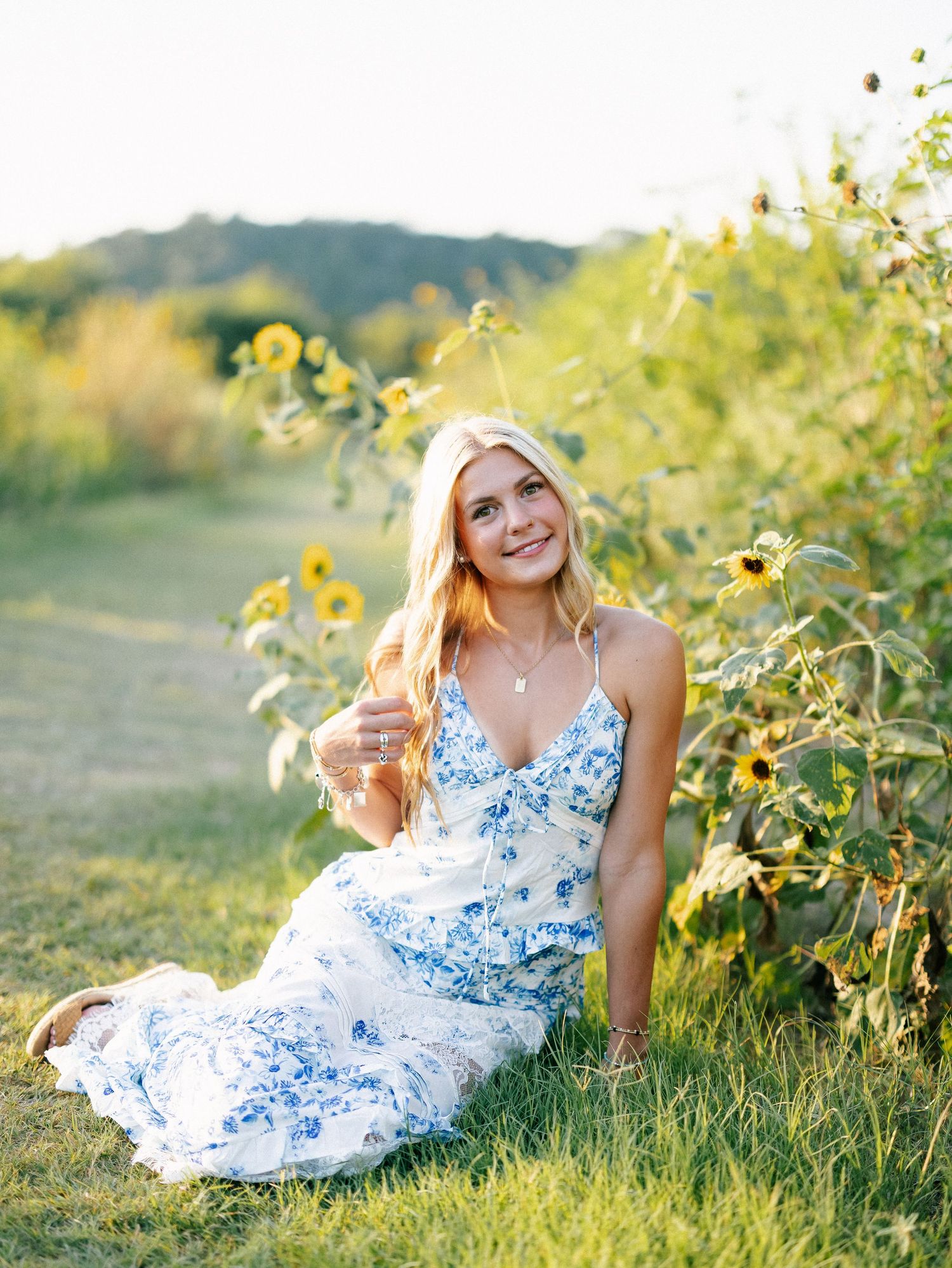 Young woman in blue floral dress sitting in sunflower field during golden hour outdoor portrait photography.