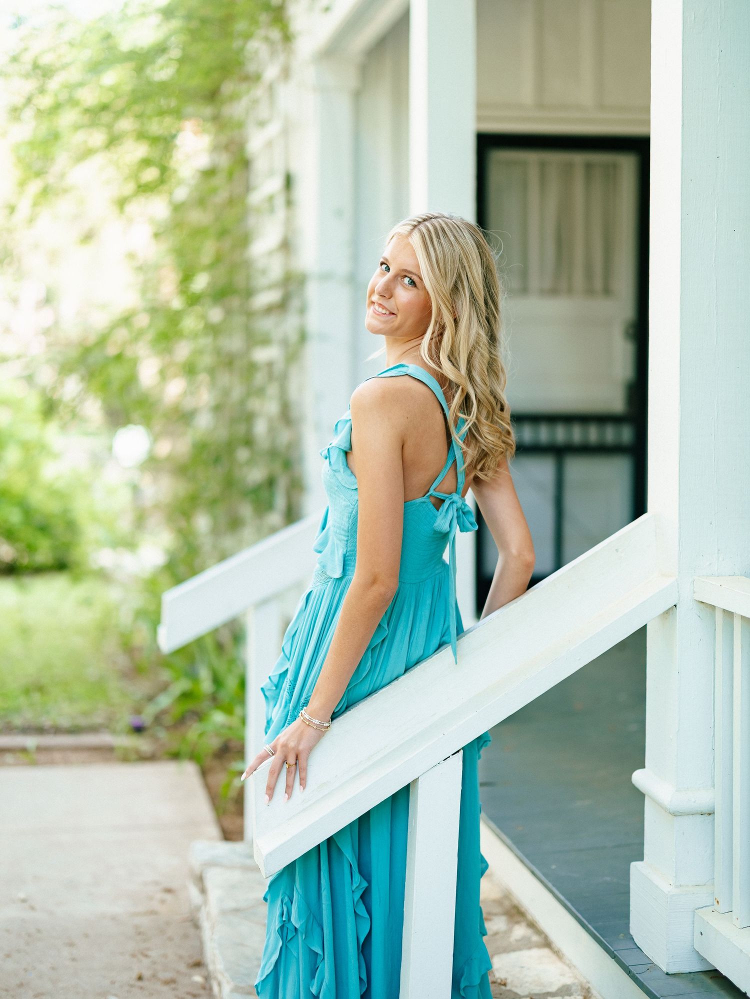 Blonde woman in teal ruffled dress posing on white porch steps surrounded by green ivy.