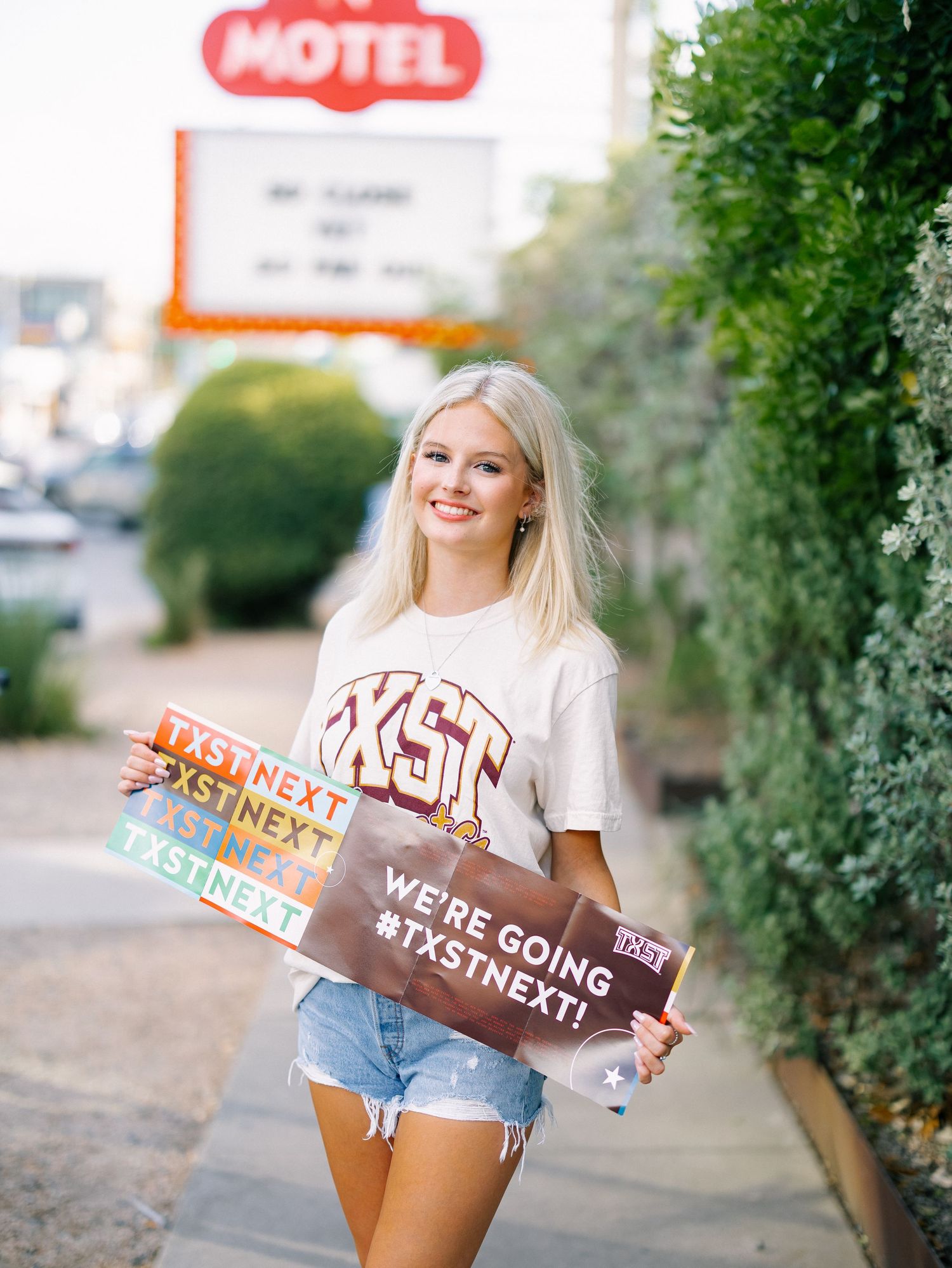 Smiling blonde teen holding TXST Next signs near motel sign, wearing white tee and denim shorts.