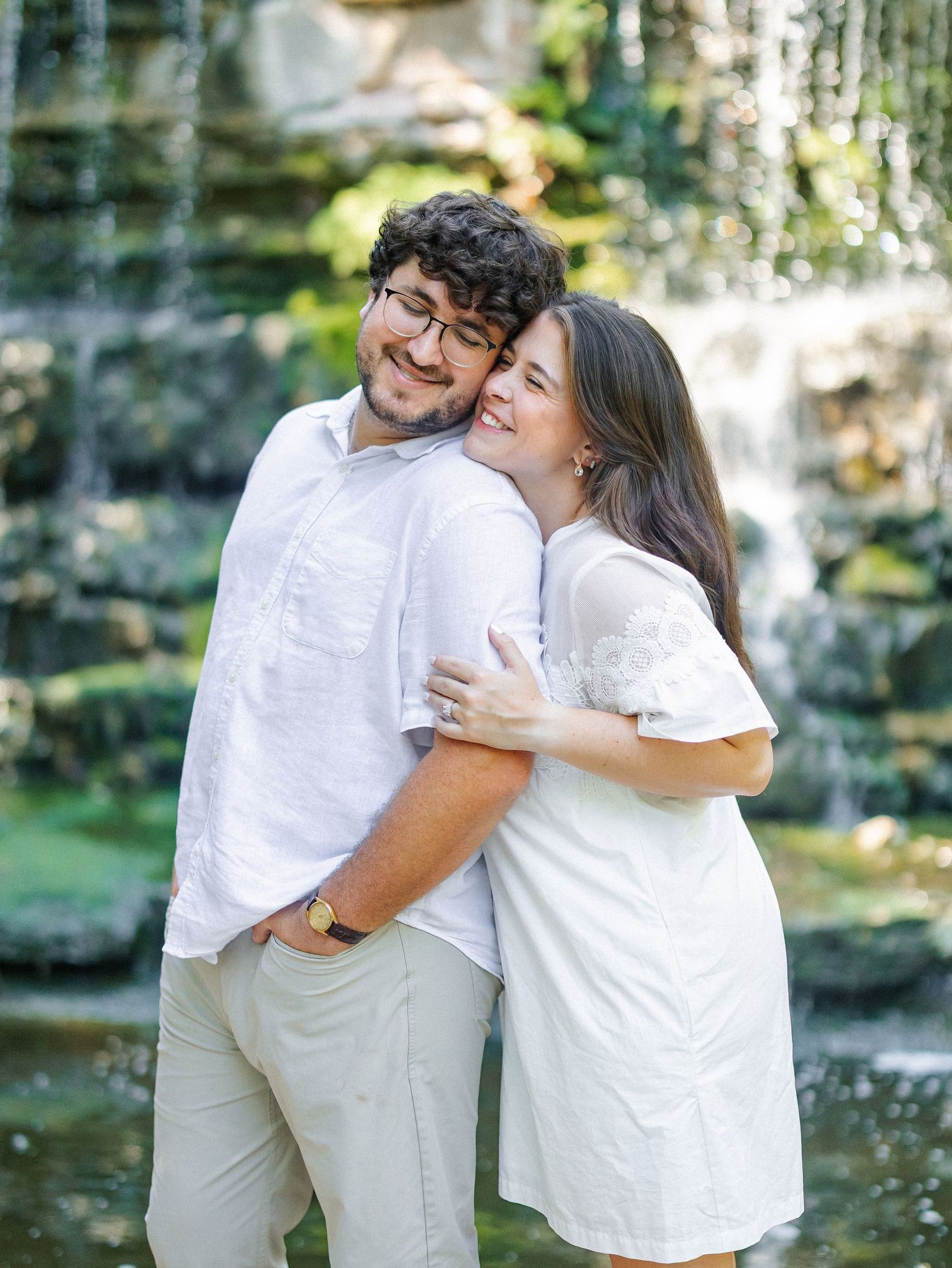 Couple in white outfits embracing and smiling in front of a waterfall in a lush garden setting.