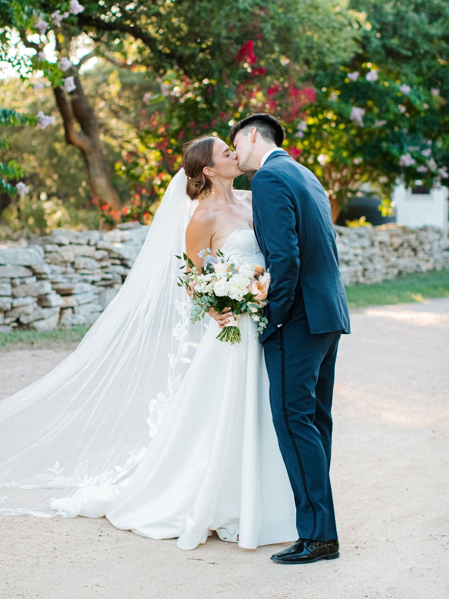 Bride in flowing gown with lace veil kisses groom in navy suit holding white floral bouquet outdoors.