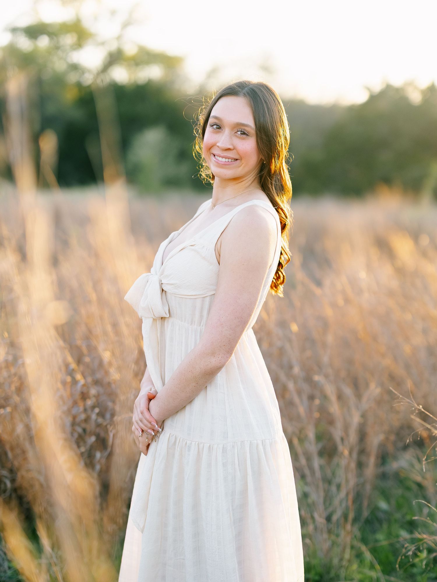 Young woman in cream sundress smiles in a golden field at sunset.