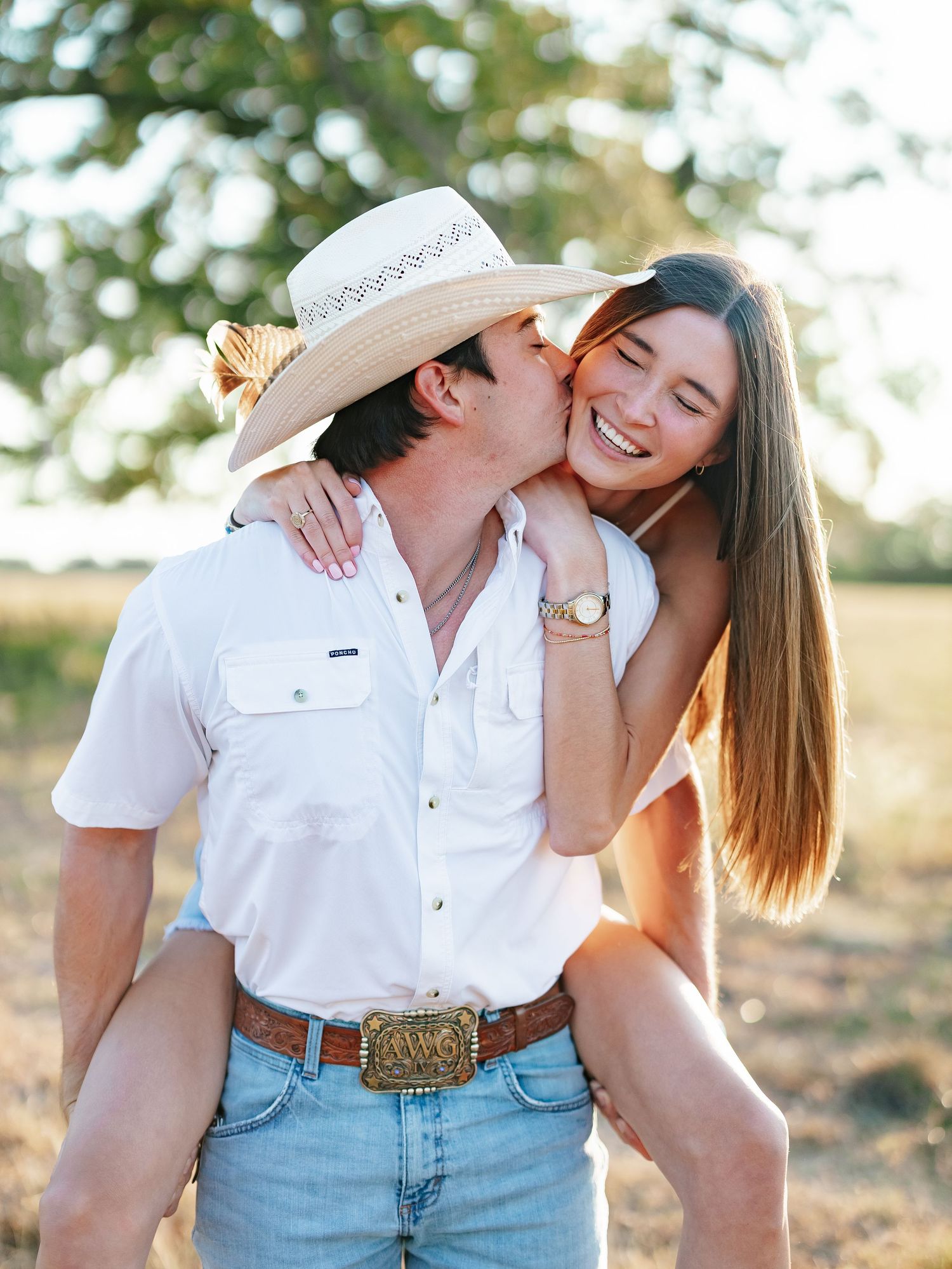 A smiling woman kisses a man in a cowboy hat on the cheek in a sunny golden field.