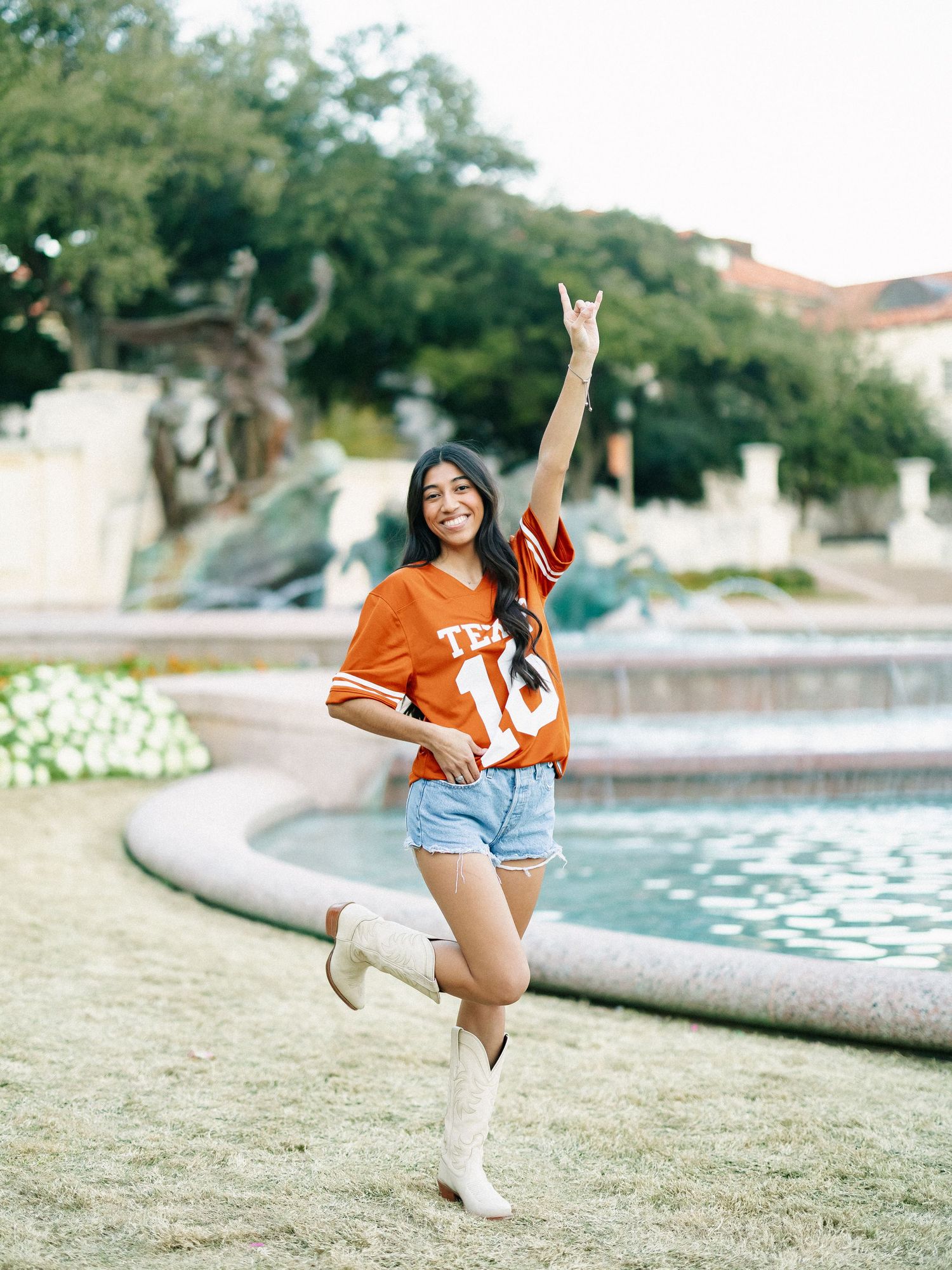 A smiling woman in a burnt orange Texas jersey, denim shorts, and white cowboy boots poses by a fountain.