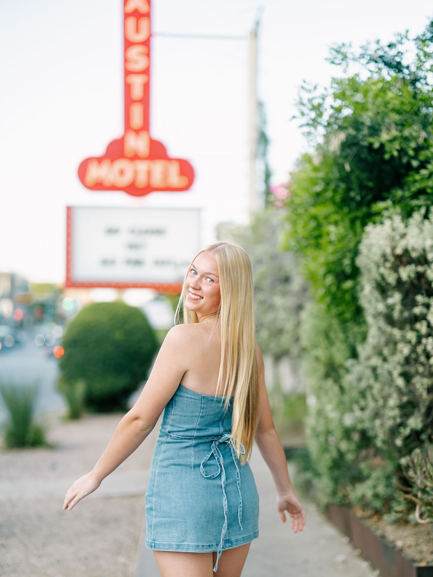 Blonde woman in denim mini dress walking near Austin Motel neon sign at dusk.