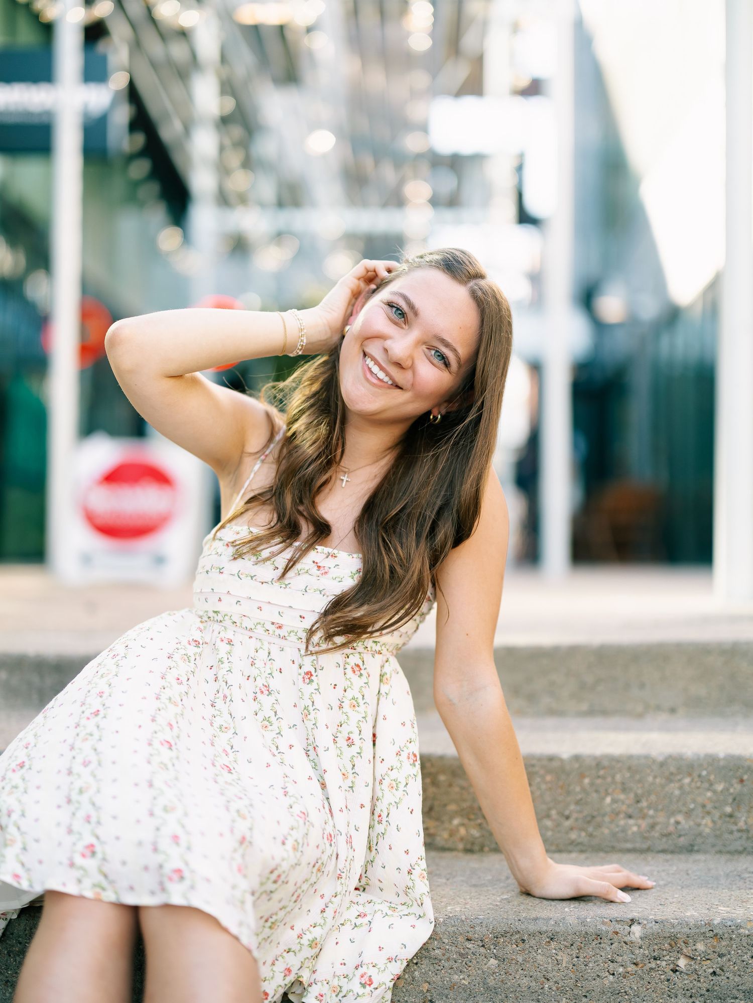 Smiling brunette in floral sundress sitting on steps with urban backdrop.