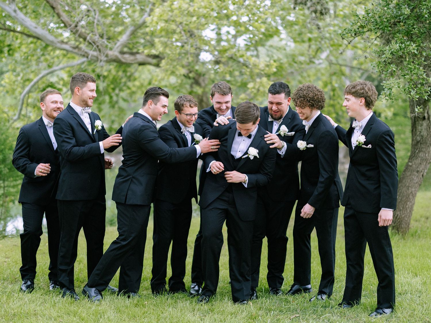 Groom and groomsmen in black suits share a fun candid moment outdoors during wedding photos.