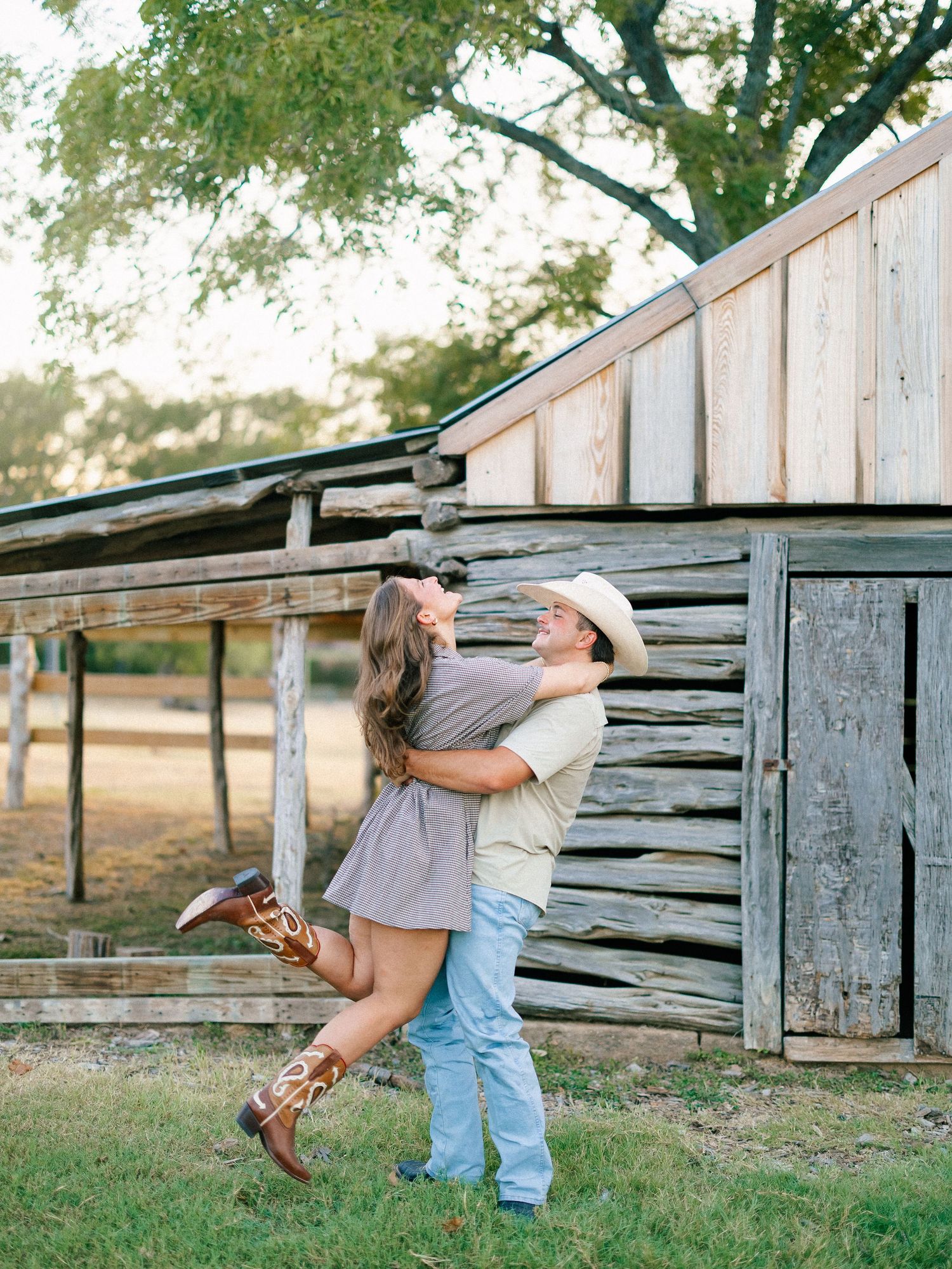 Man lifting woman in an embrace near a weathered wooden barn during a romantic country engagement session.