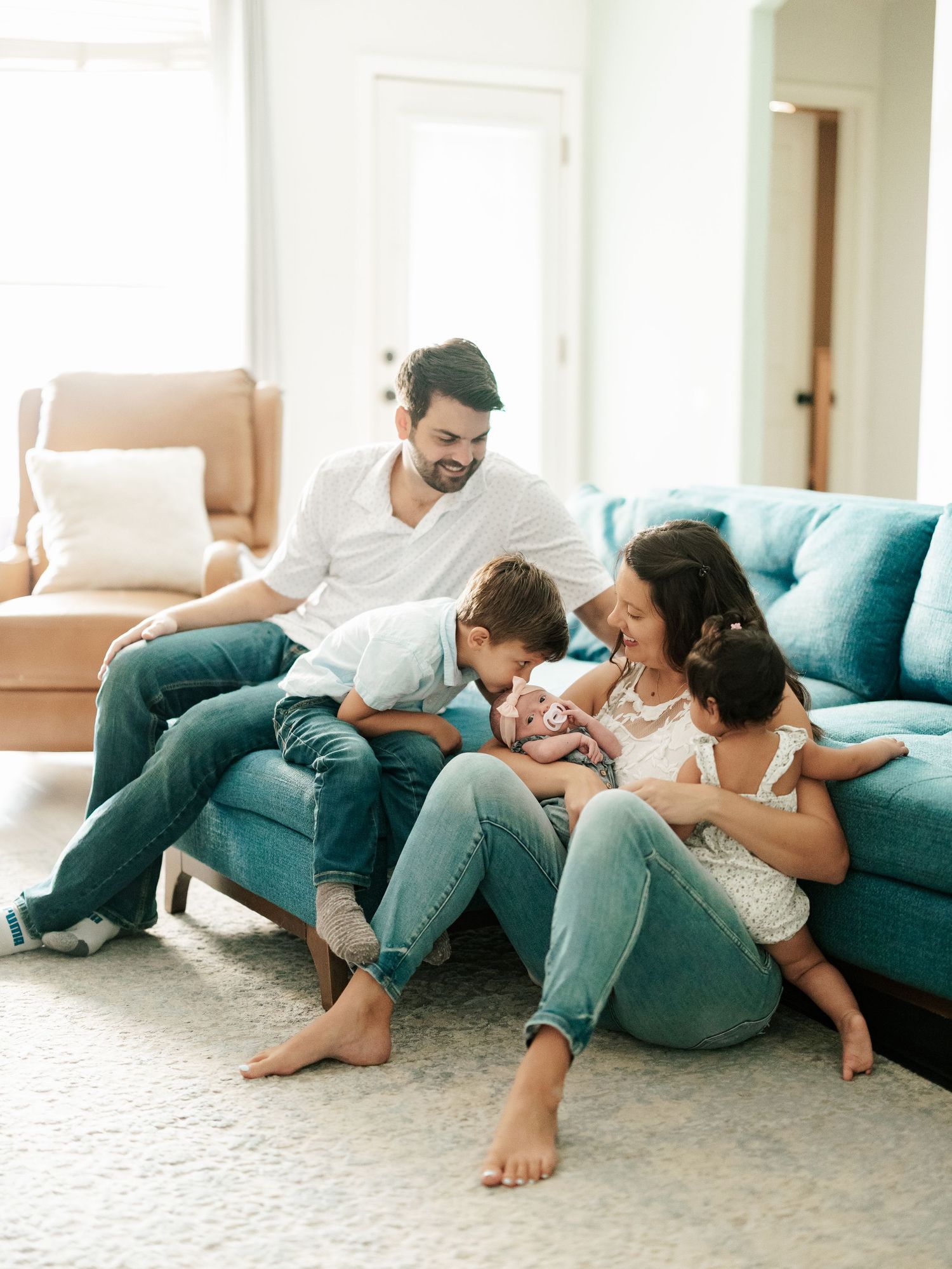 Family of five sitting on teal sofa, parents smiling at newborn baby while siblings gather close in bright living room.