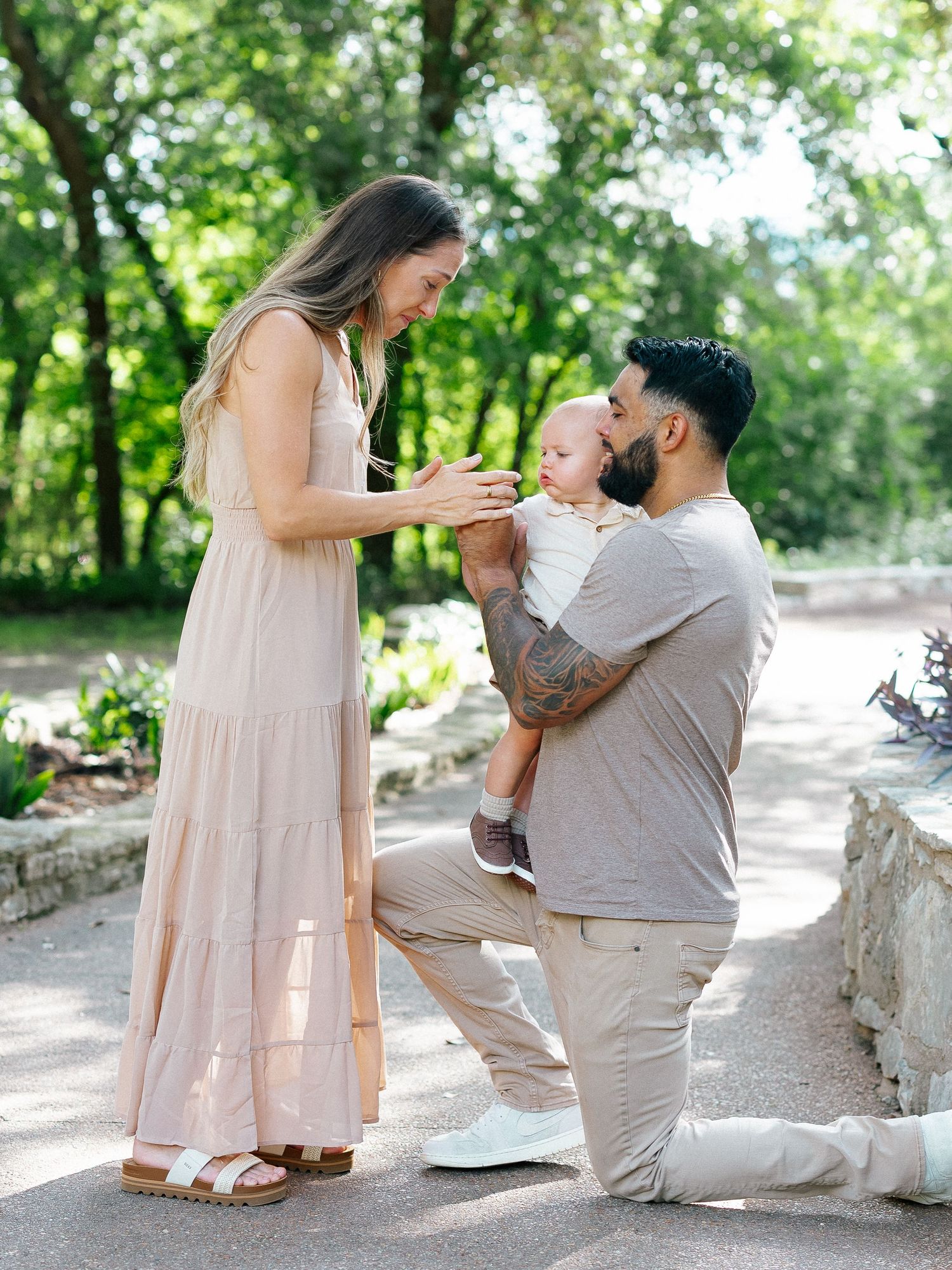 A man kneels on one knee holding a baby while a woman reaches out in a sunny park.