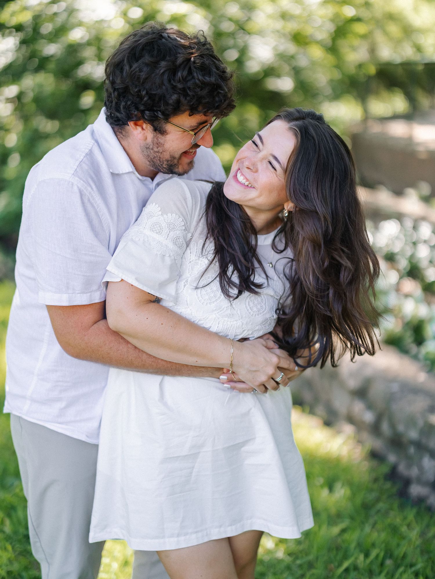 A happy couple in matching white outfits embrace and laugh together outdoors in a lush green garden setting.