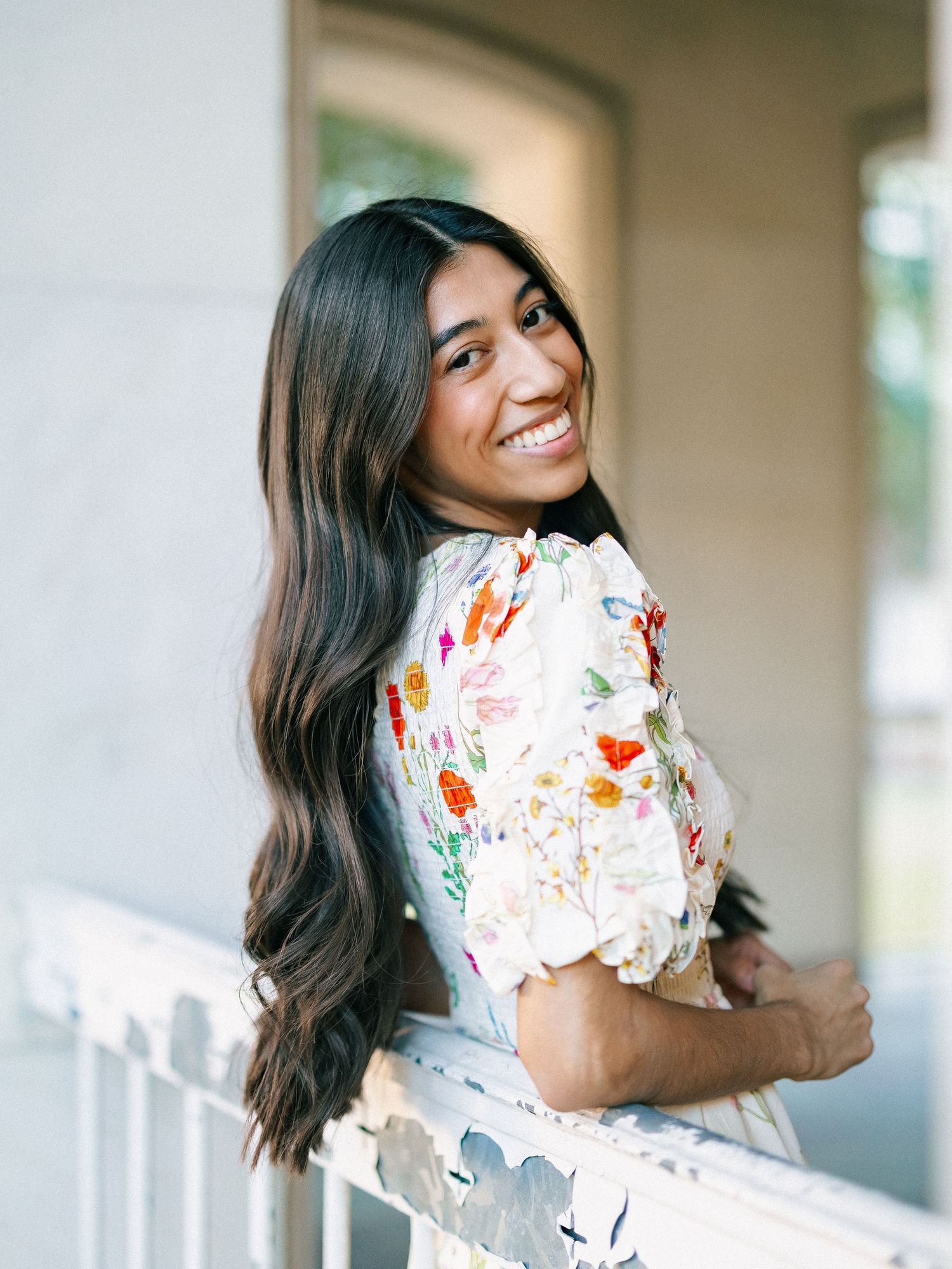 A smiling young woman with long dark hair wearing a floral dress leans against a white railing outdoors.